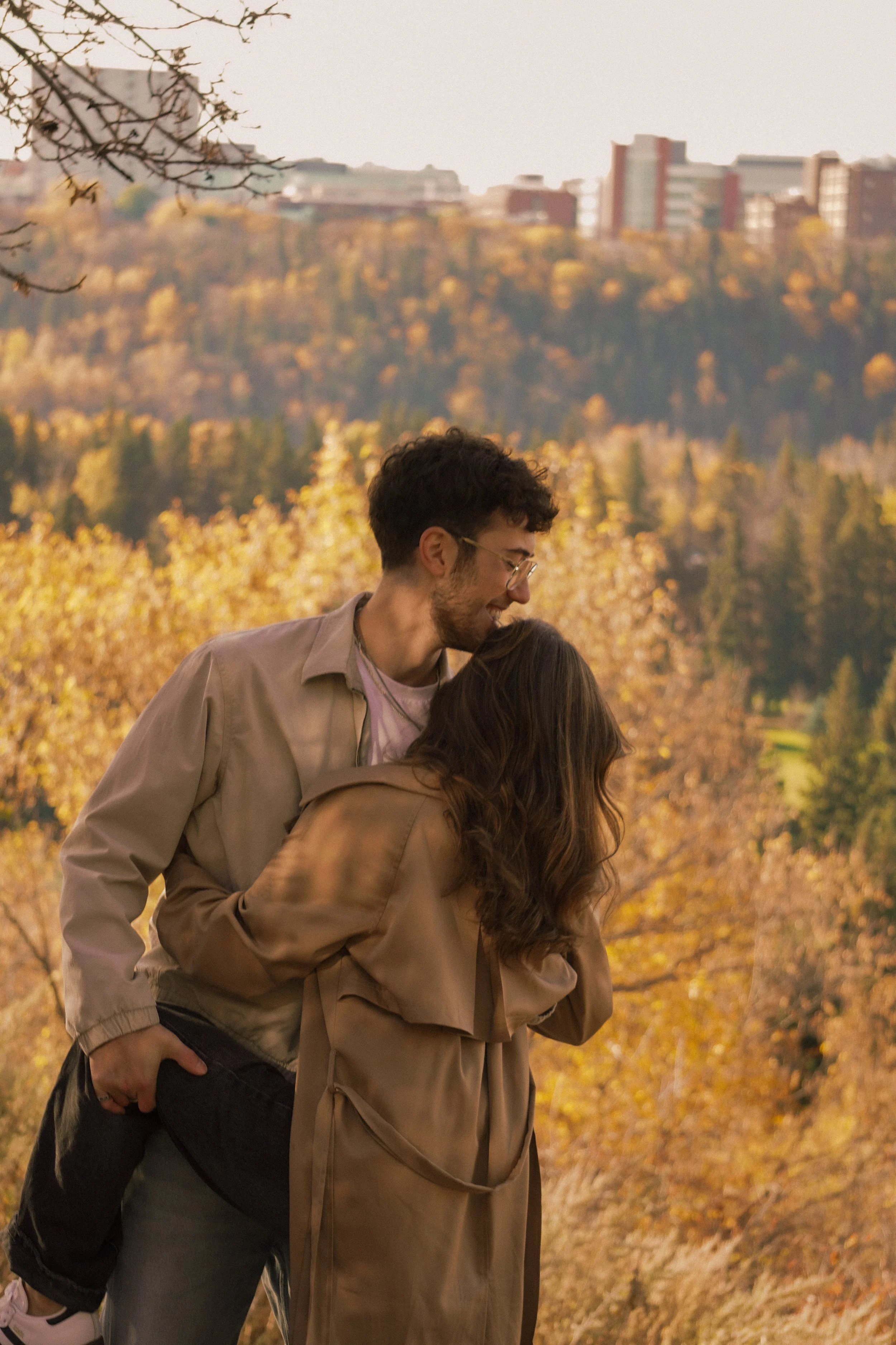 A couple embracing outdoors during autumn, surrounded by trees with fall foliage and city buildings in the background.