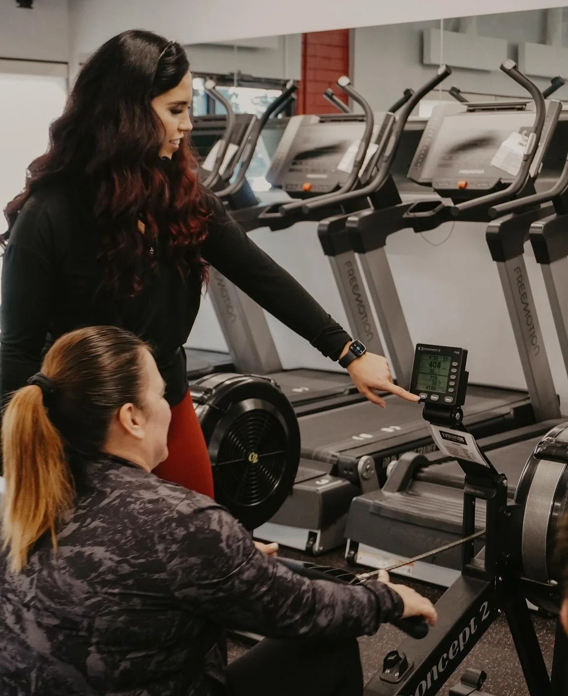 Two women in a gym next to treadmills. One woman is sitting on an exercise bike, while the other points at the bike's digital display, possibly explaining or adjusting workout settings.