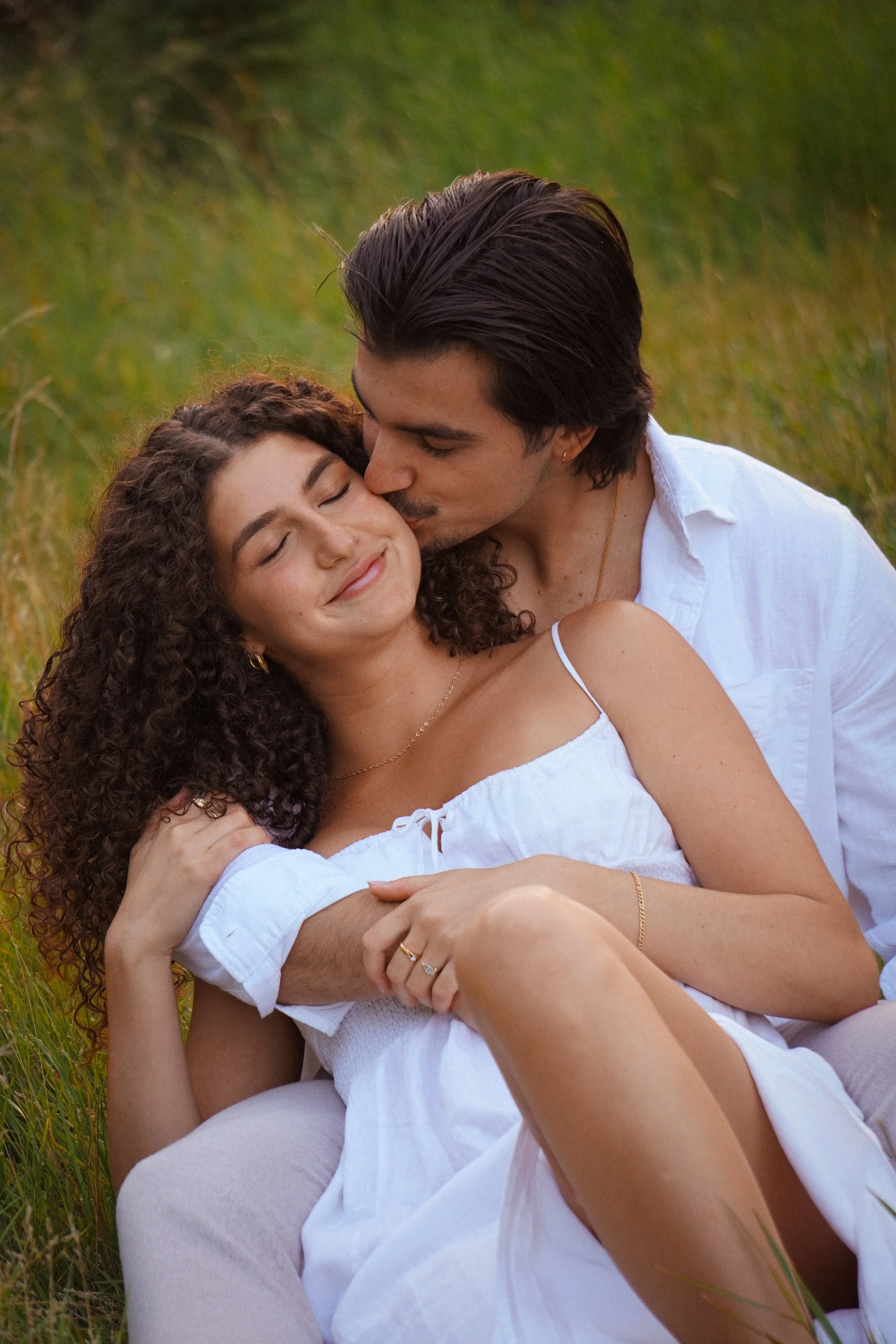 A young couple sitting outdoors on grass, with the man kissing the woman on her cheek. The woman has curly hair and is smiling with her eyes closed, wearing a white dress. The man has dark hair, is wearing a white shirt, and has his arms around the woman.