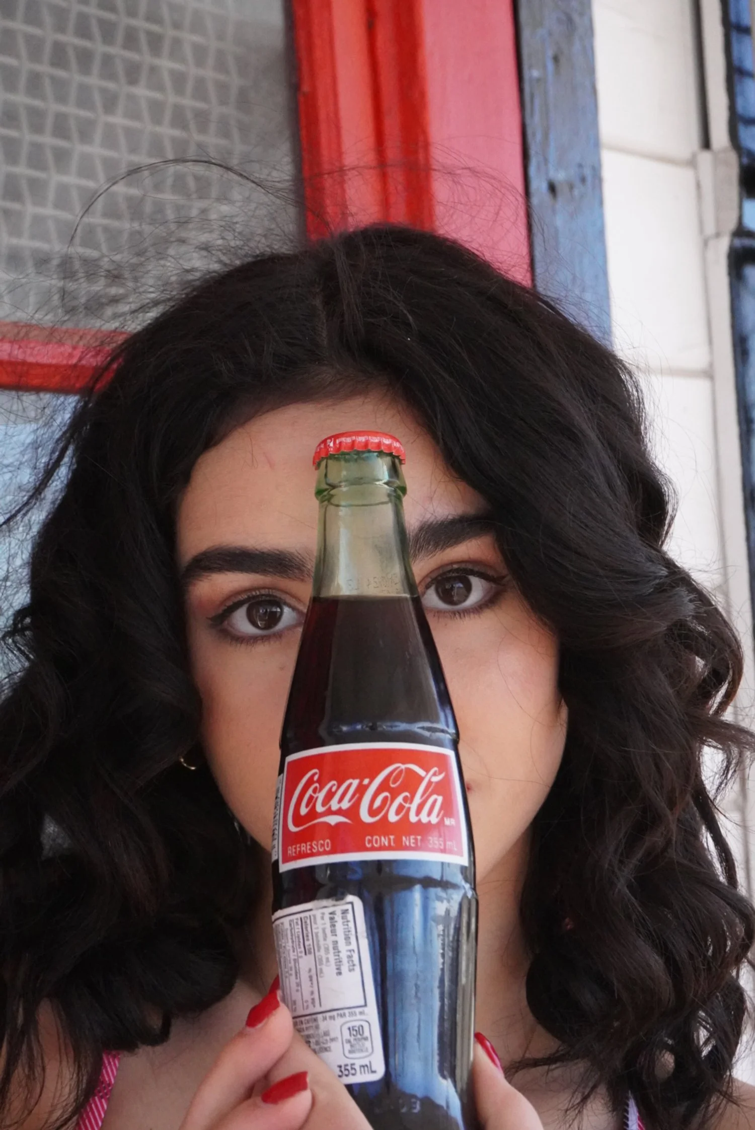 Young woman with curly dark hair and red nail polish holding a Coca-Cola bottle up to her face, with her eyes visible behind the bottle.