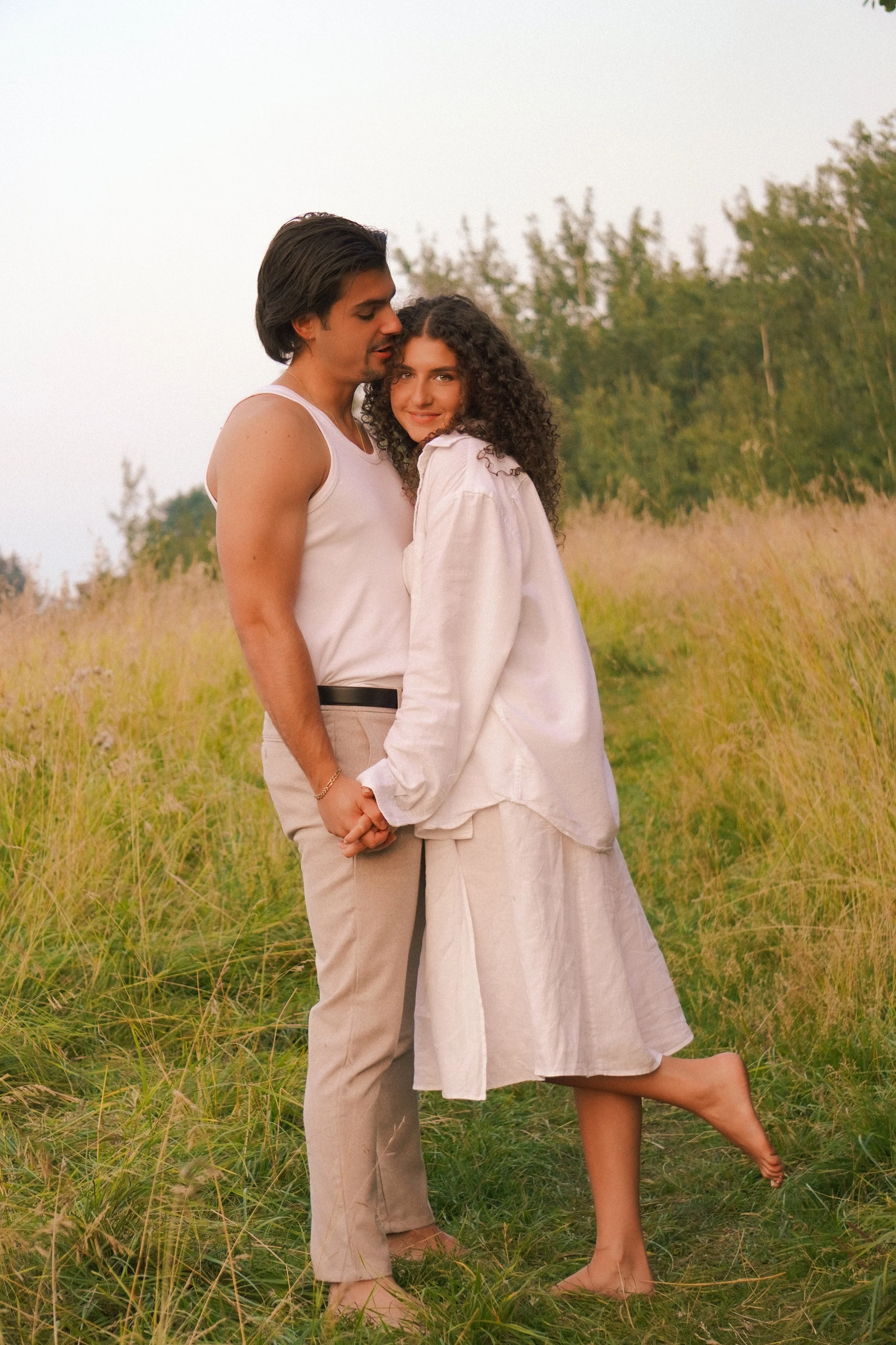 A couple standing in a grassy field holding hands, with trees in the background during sunset.