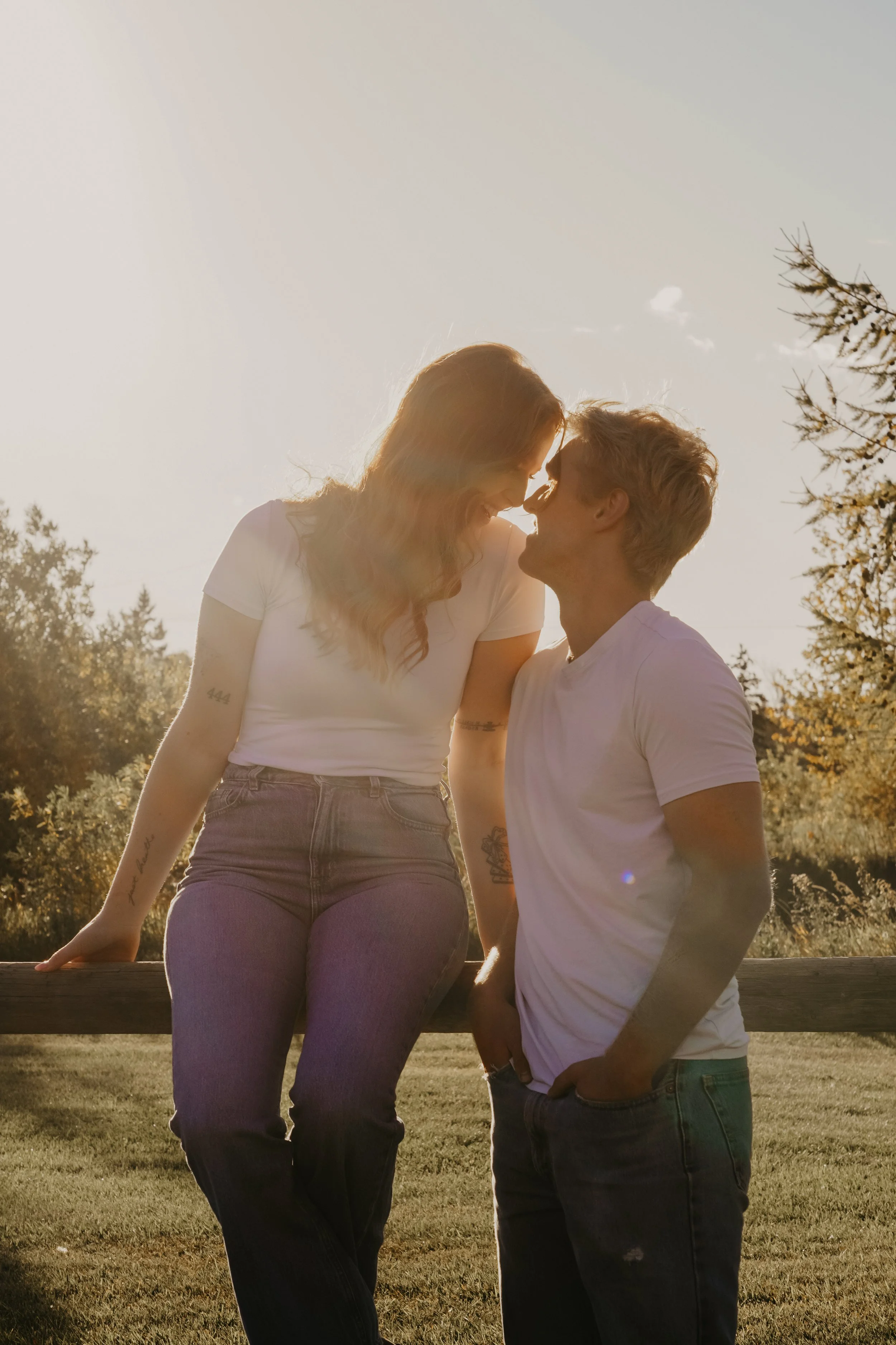 A couple is outside at sunset, smiling and leaning towards each other, with the woman sitting on a wooden railing and the man standing beside her, surrounded by trees and grass.