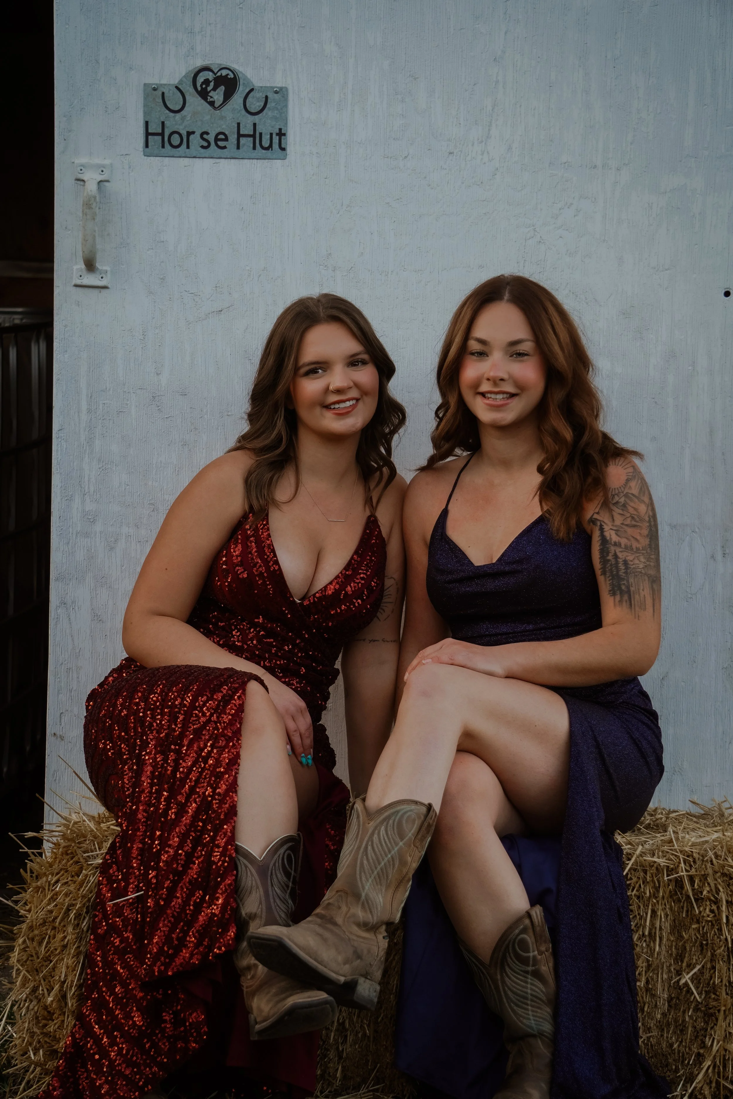 Two young women sitting on a hay bale in front of a white wall with a sign that reads 'Horse Hut' and a horse logo. They are wearing dresses and cowboy boots, smiling at the camera.