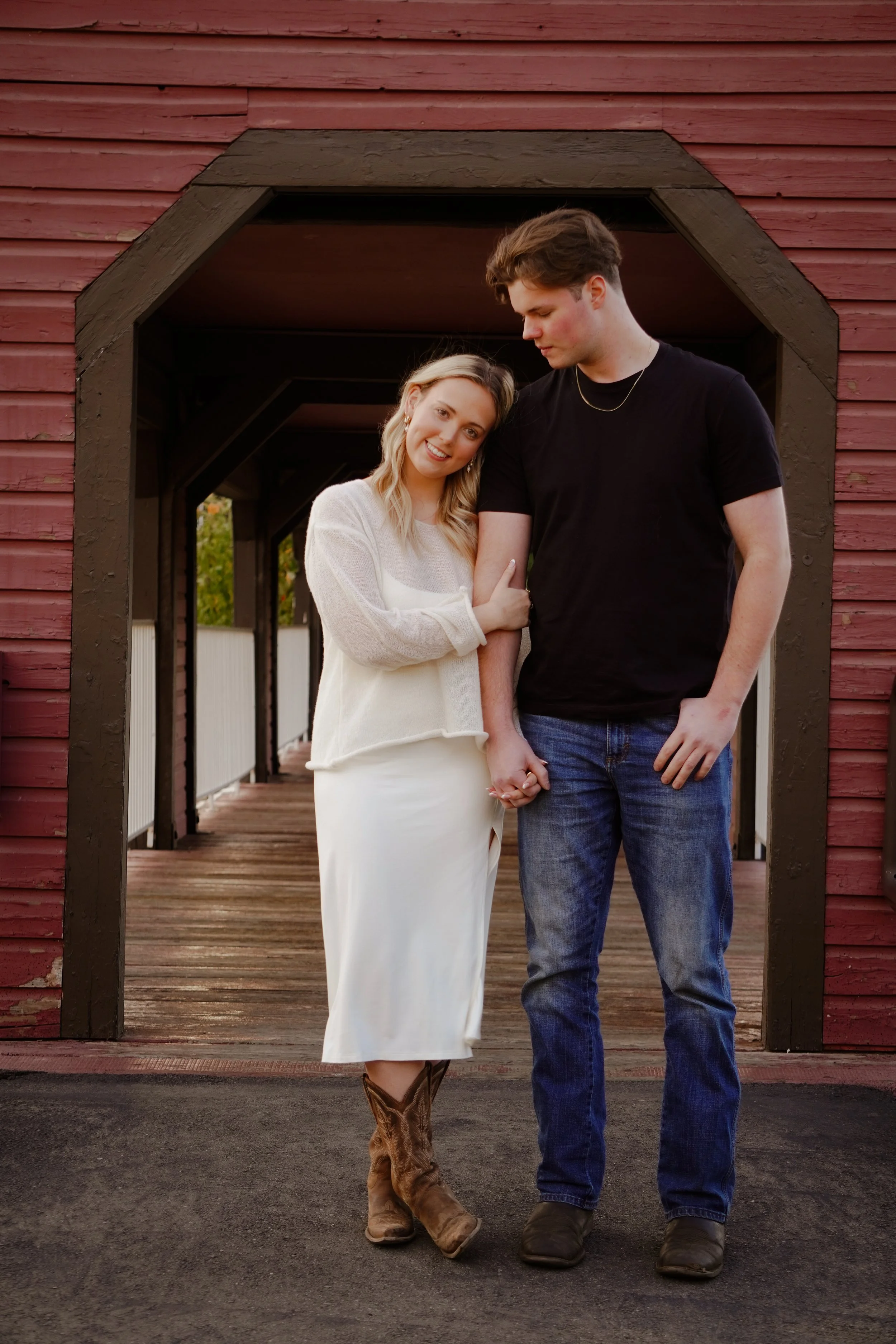 A young couple holding hands and smiling while walking on a wooden bridge with a red barn in the background.