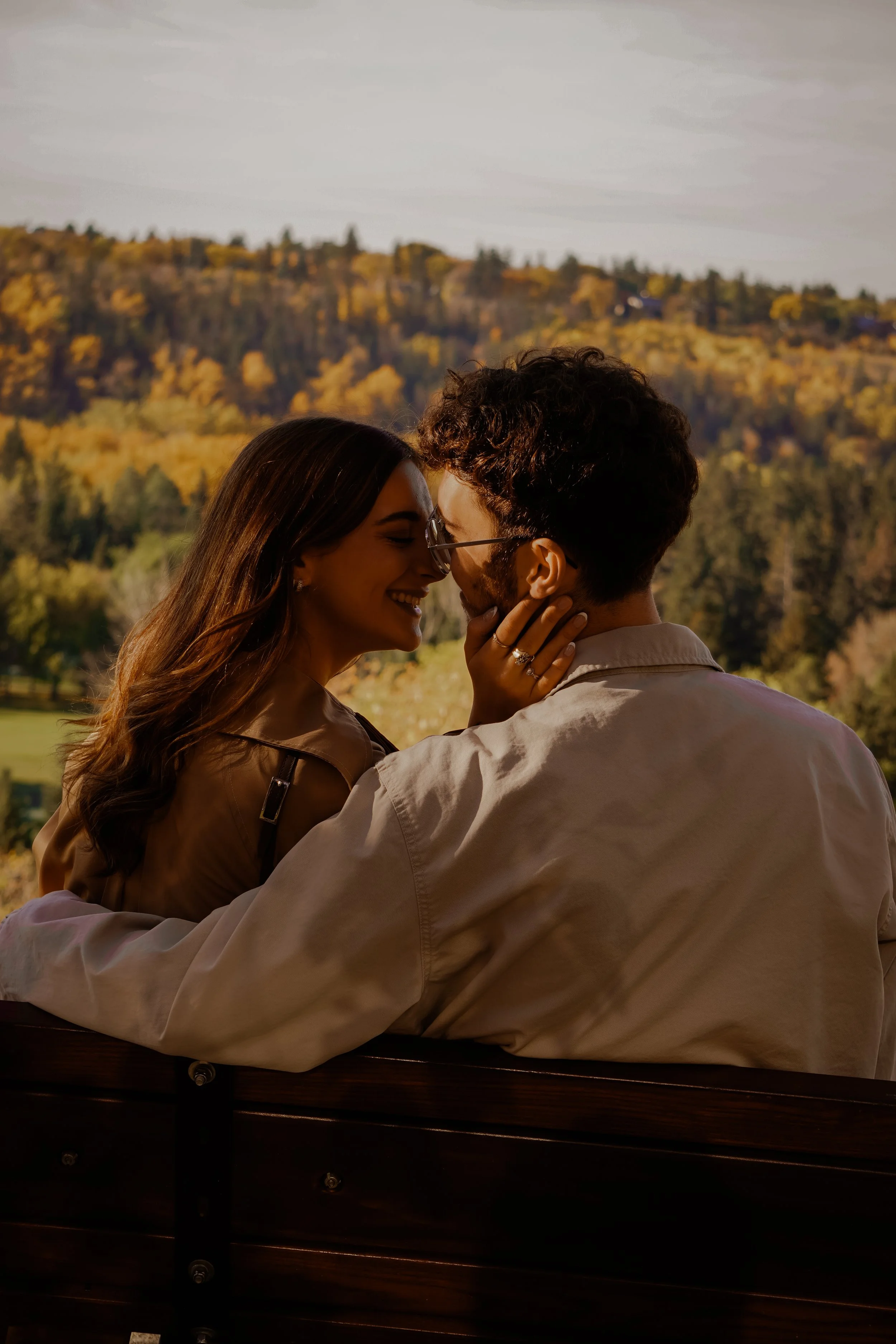 A couple sitting on a park bench, leaning close and smiling at each other, with a scenic backdrop of autumn trees and hills.