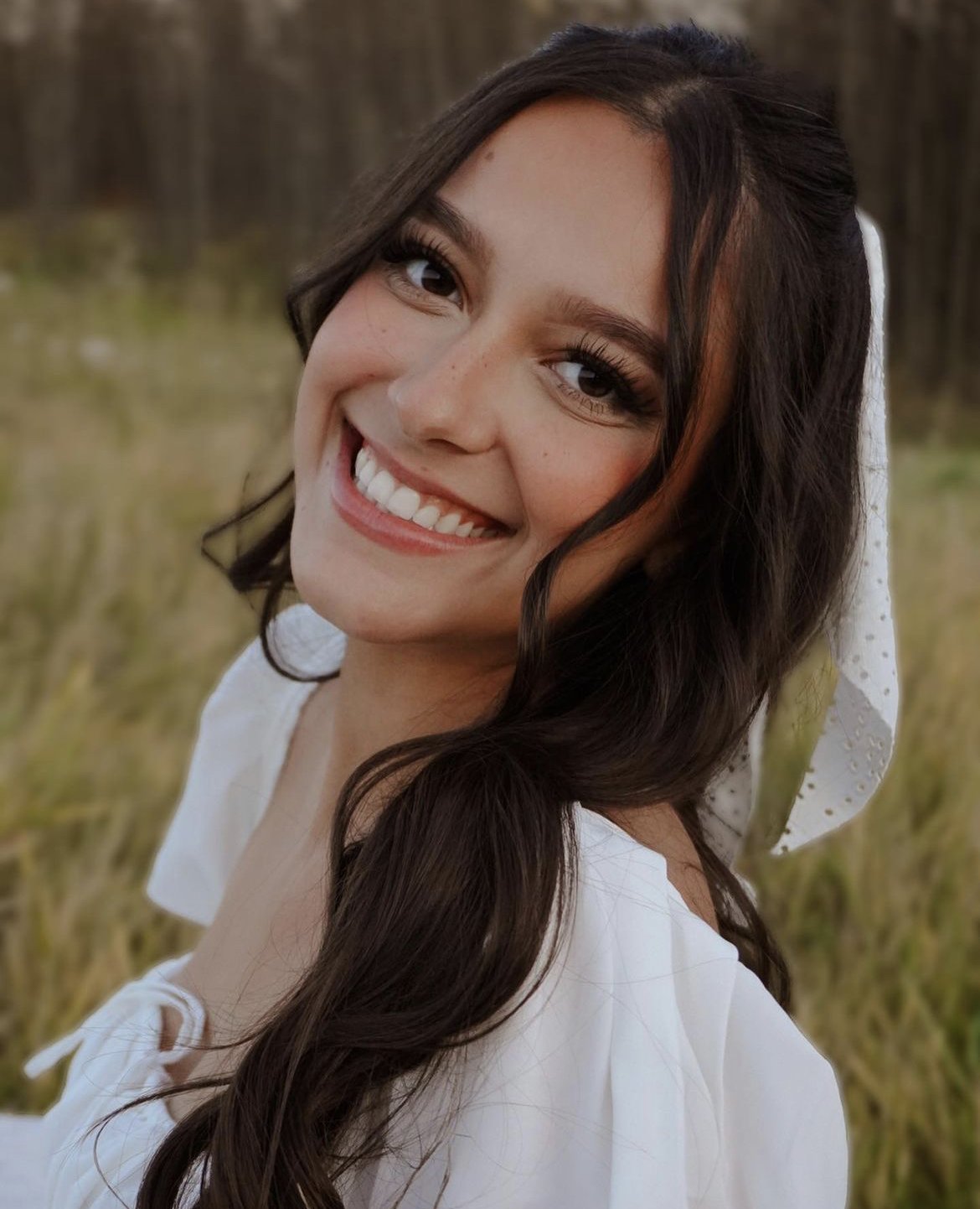 Close-up of a young woman with long dark hair, smiling, outdoors in a grassy field with trees in the background, wearing a white top and a white headband.