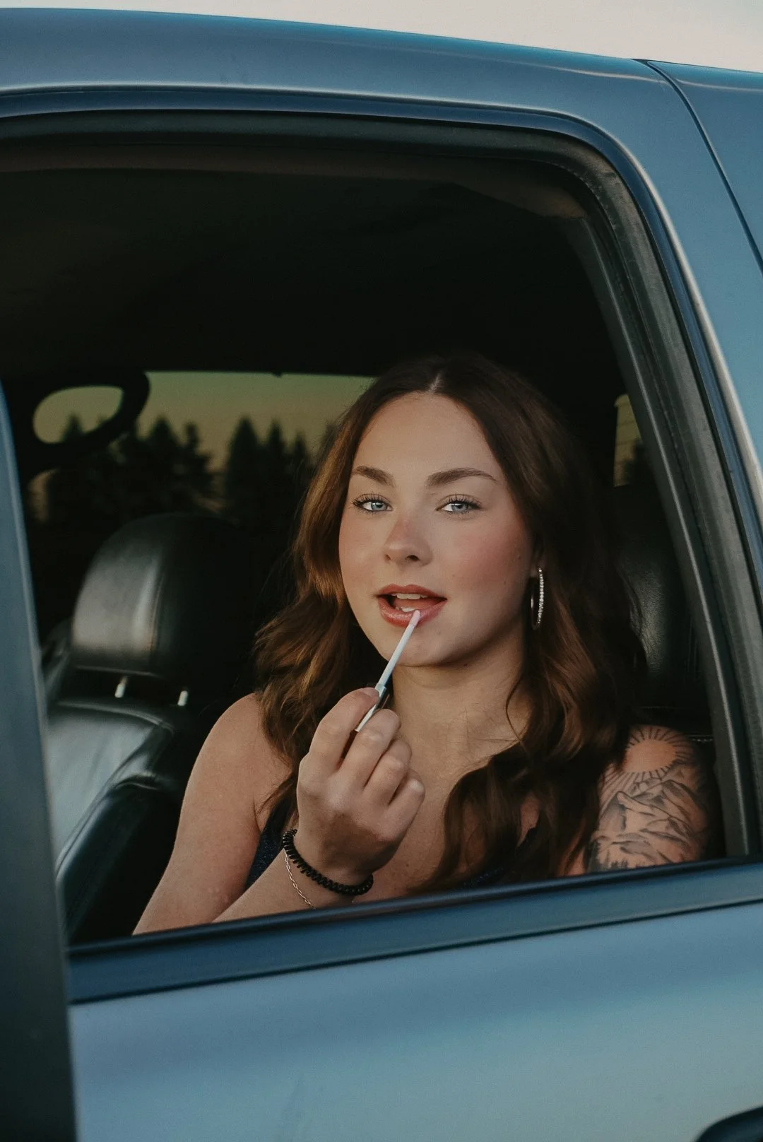 A young woman with wavy brown hair, blue eyes, and hoop earrings sitting in a car, applying lipstick with a lip brush, and looking at the camera.