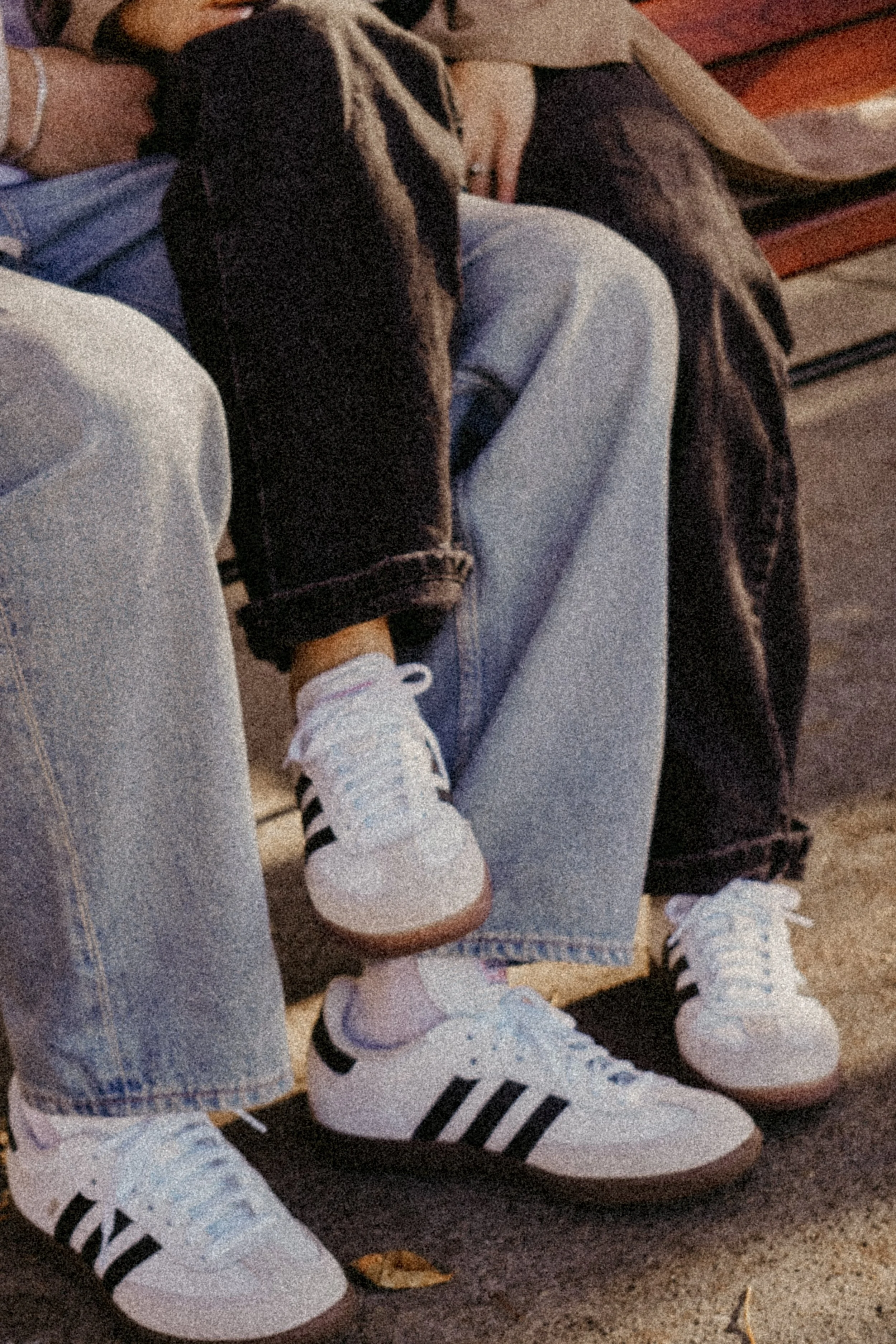 Two people sitting on a bench, wearing light blue jeans and white sneakers with black stripes, and a dog with a black coat and white markings resting one paw on the person's leg.