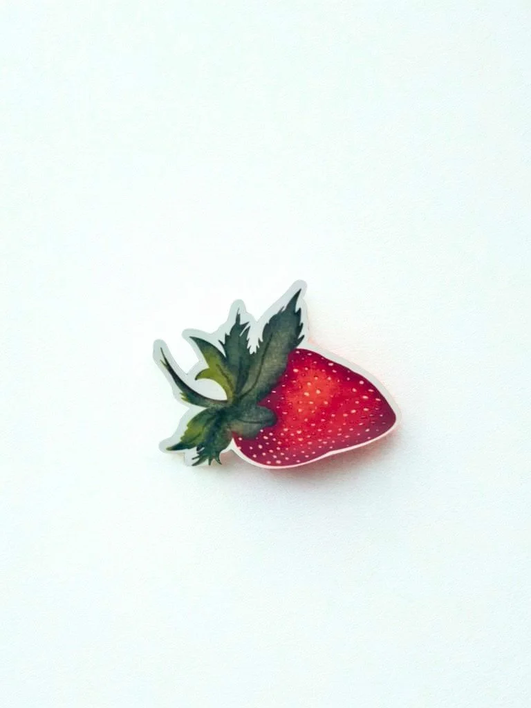 Hand-illustrated ripe red strawberry die-cut sticker with leafy green top displayed on a white background.