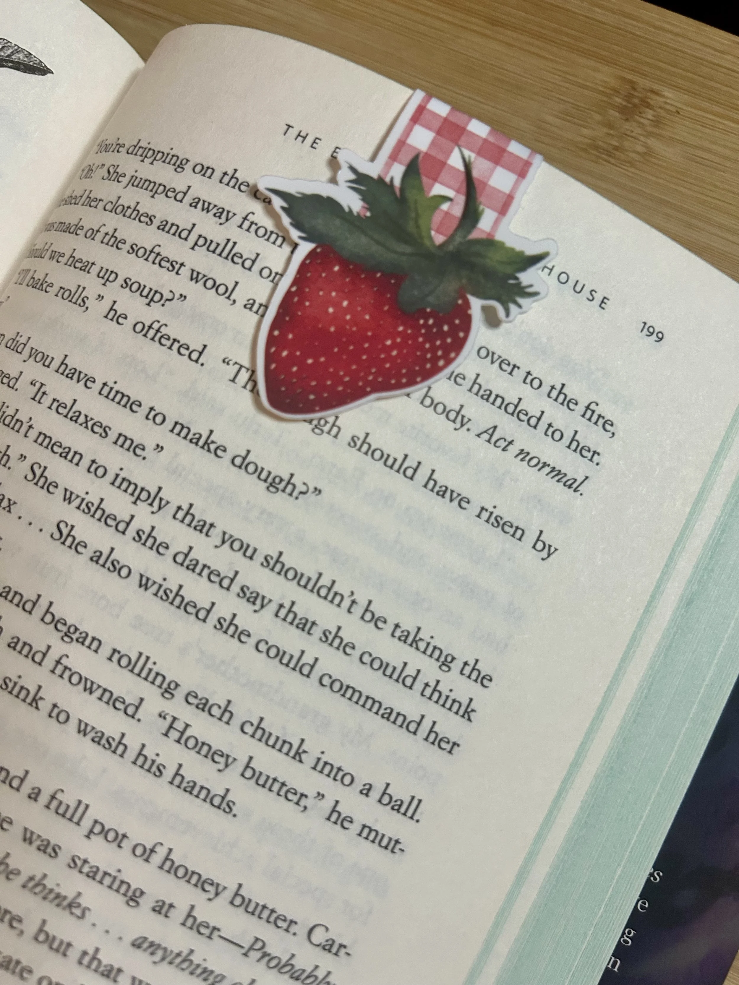 Handcrafted strawberry magnetic bookmark with hand-illustrated red strawberry and pink gingham tab from Gather’s Strawberry Summer Collection shown in position on a paperback novel