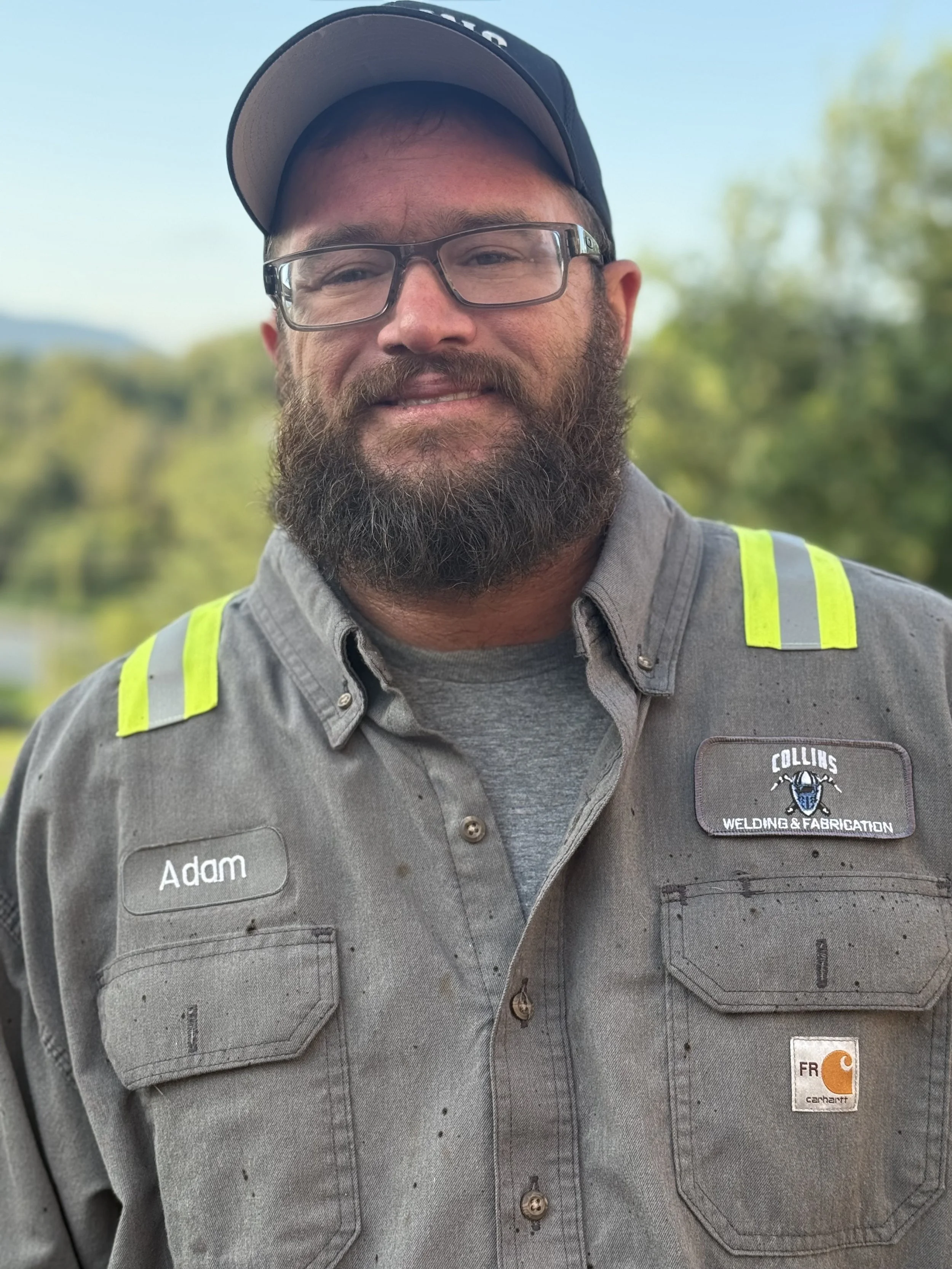 Close-up of a man wearing safety glasses, a cap, and a work shirt with reflective stripes, smiling outdoors against a background of trees and blue sky.