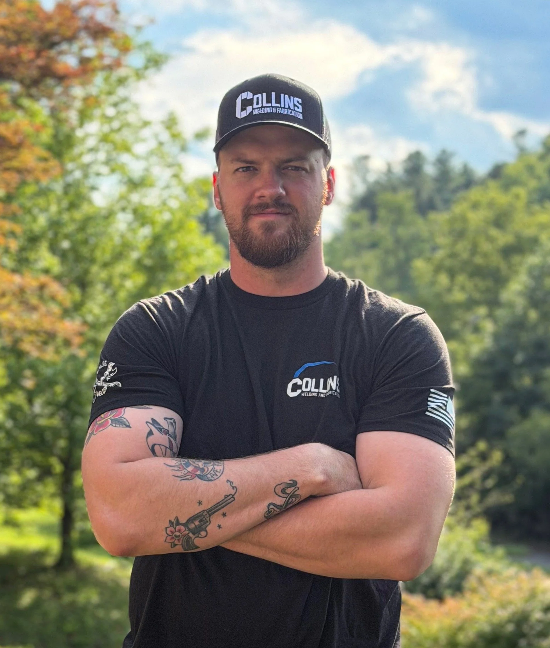 A man with a beard and tattoos on his arms wearing a black cap and black t-shirt, standing outdoors with crossed arms, trees and a cloudy sky in the background.