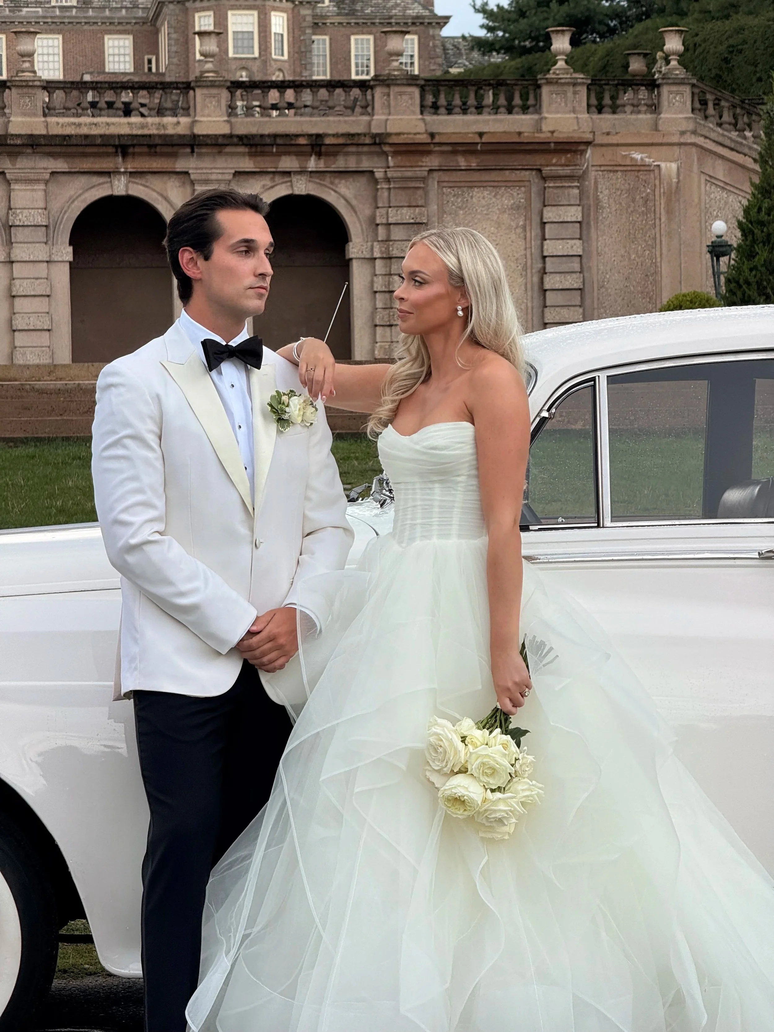 A bride and groom in wedding attire standing near a white vintage car in front of a historic building, with the bride holding a bouquet of white roses.