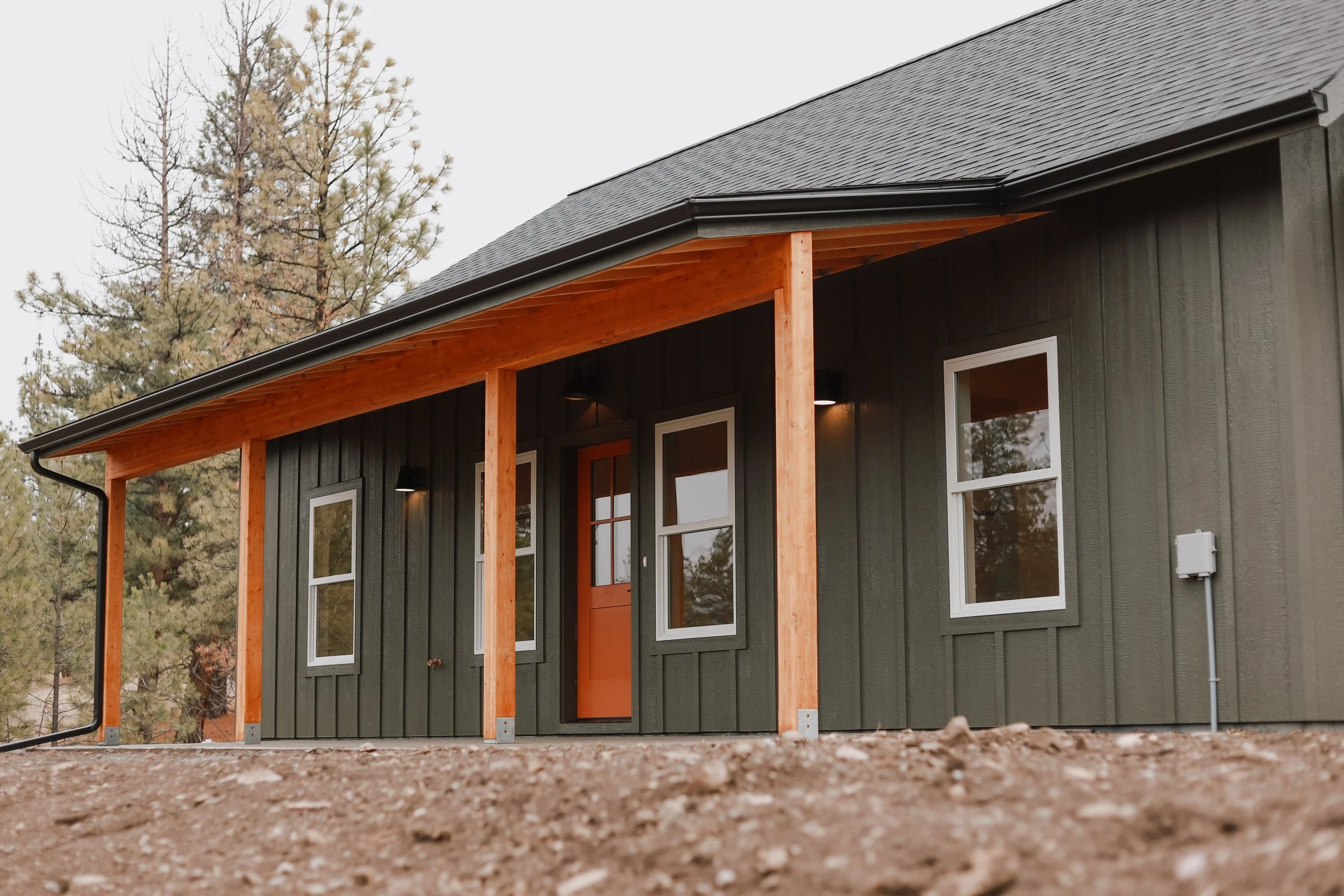 This photo shows a newly built modern-rustic home in Hot Springs, MT, featuring green board-and-batten siding, warm wood porch posts, and a striking orange front door set against a backdrop of tall evergreens.