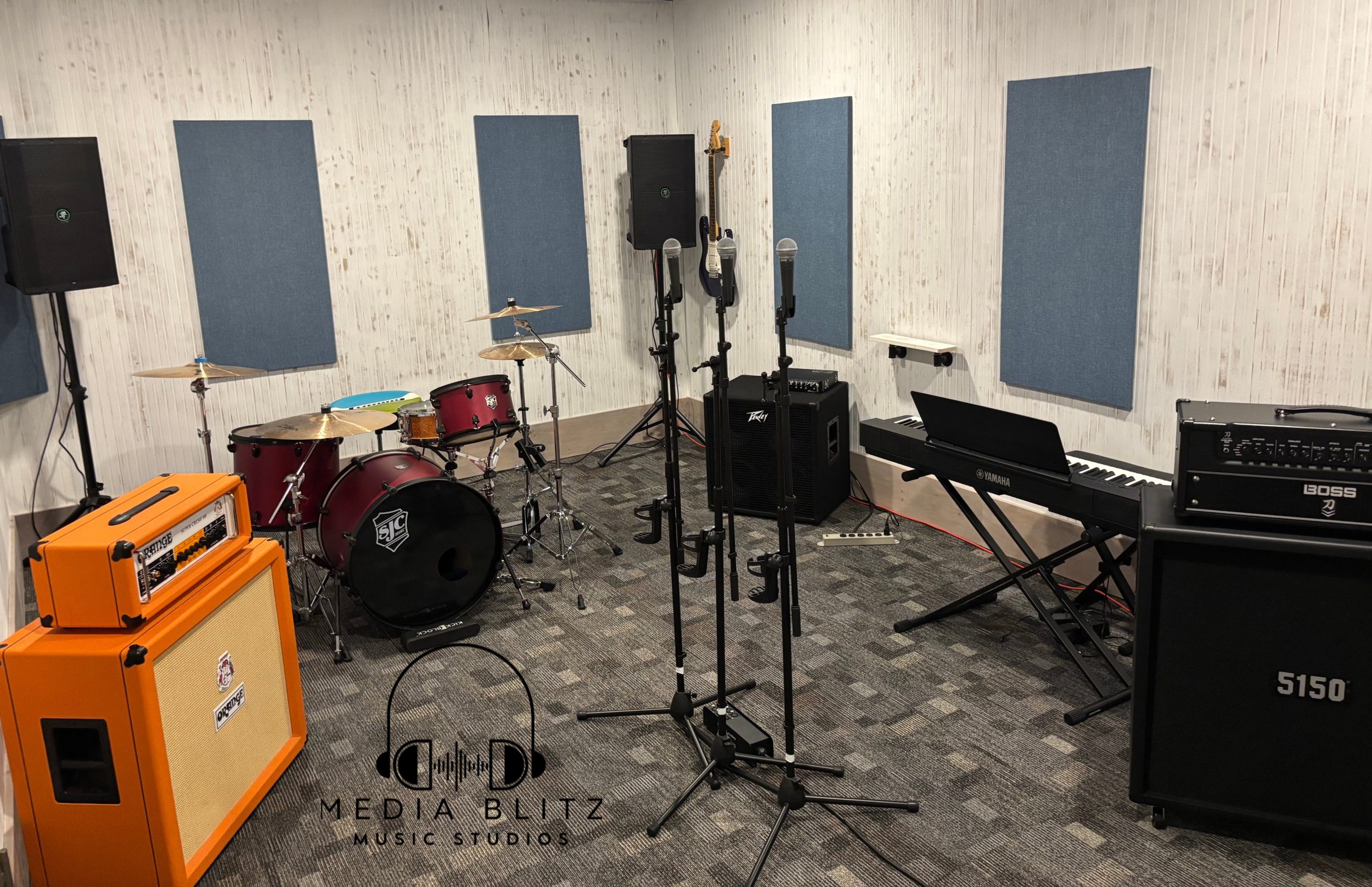 A music rehearsal room with various instruments and equipment, including a drum set, keyboards, amplifiers, microphones, and acoustic panels on the walls.