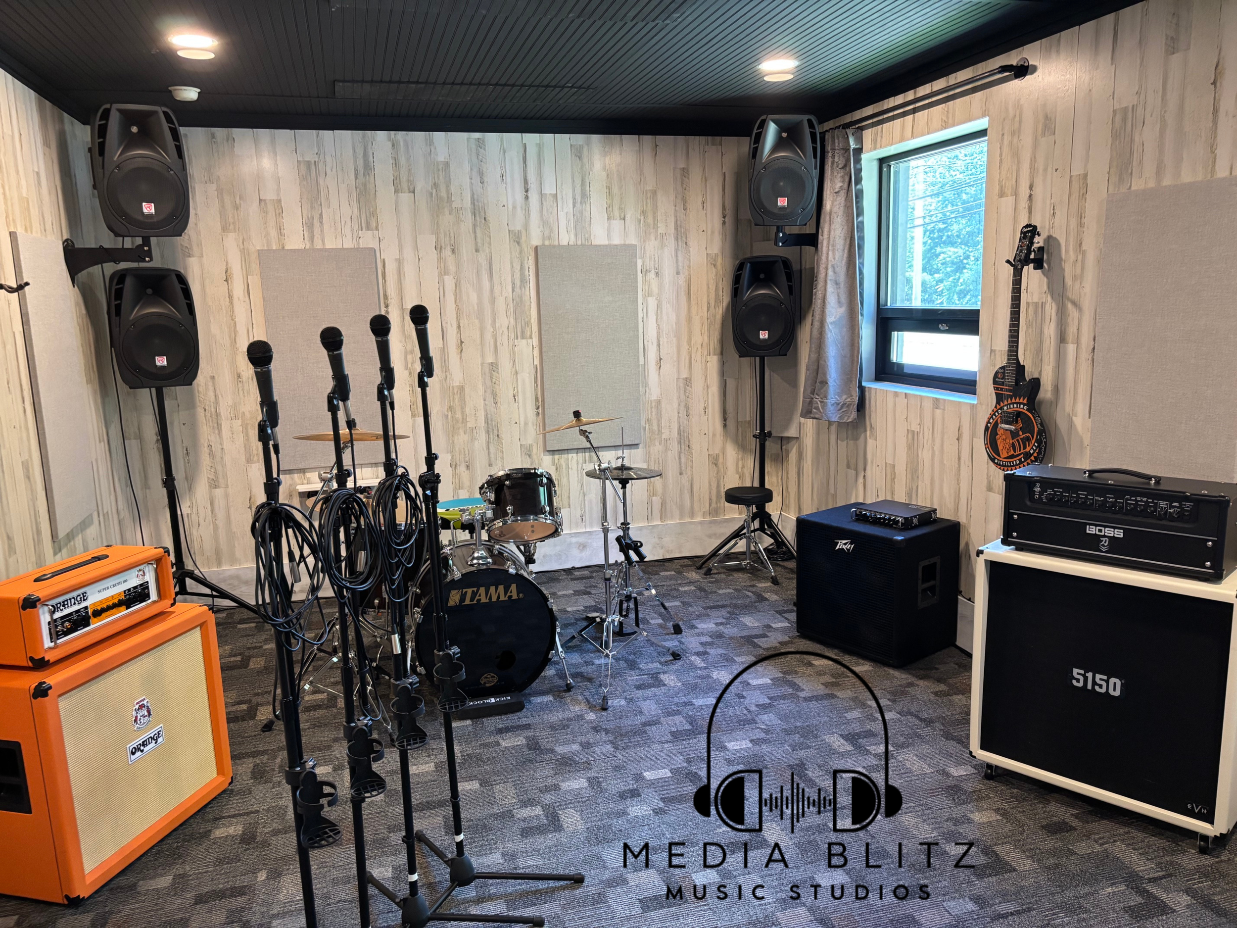 Music rehearsal room with drums, microphones, amplifiers, speakers, and a guitar hanging on the wall near a window.