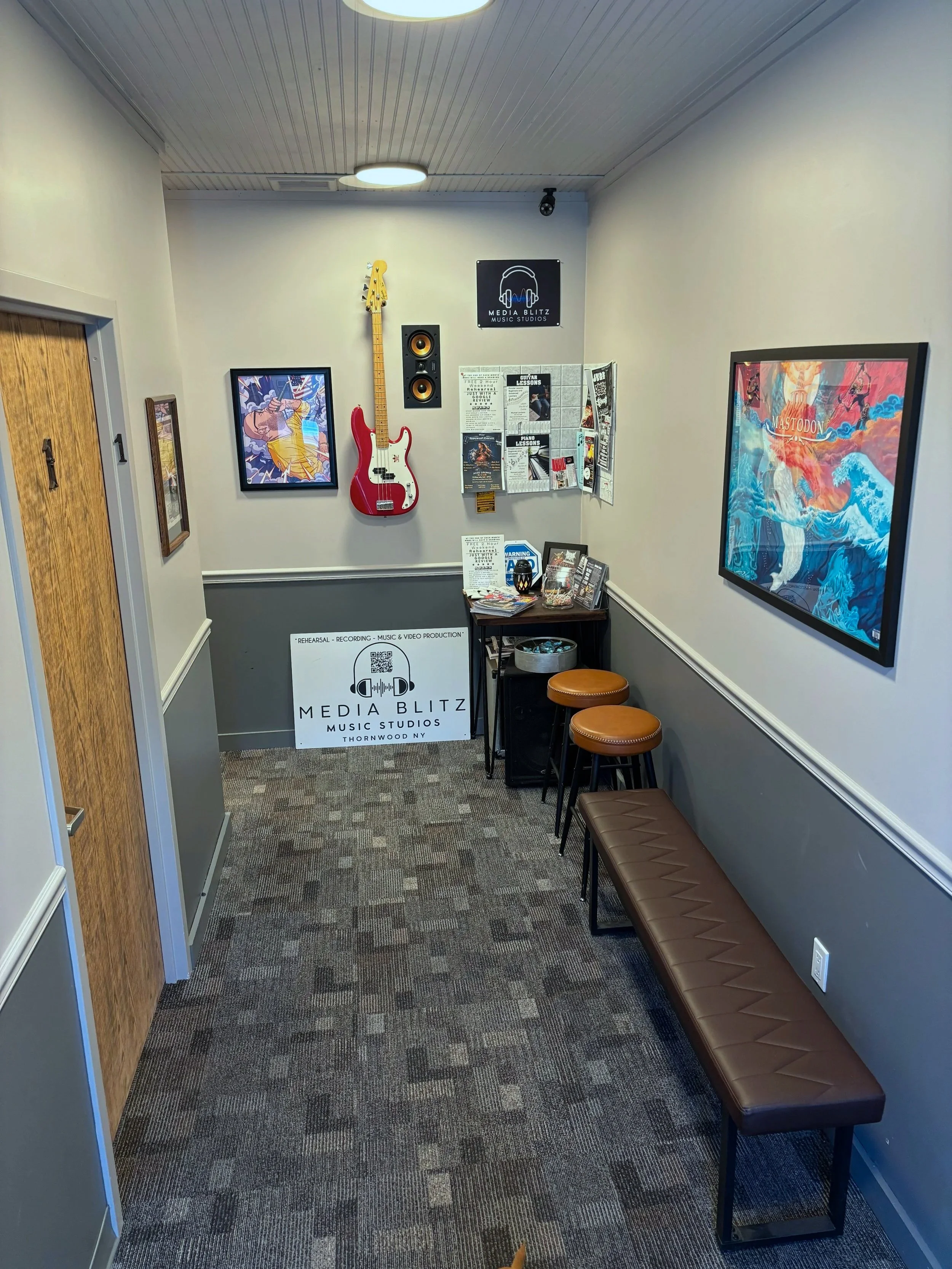 A small music studio with a framed Mastodon poster on the wall, a guitar hanging next to a speaker, a media blitz sign at the back, a small table with magazines and a sign, and a long brown bench along the right wall.
