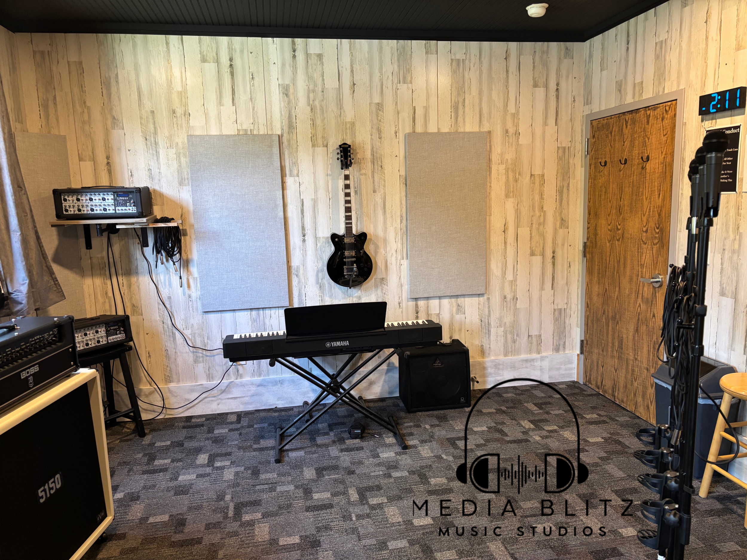 A music studio with a Yamaha keyboard, guitars, sound equipment, and acoustic panels on a wood-paneled wall, with a digital clock showing 2:11.