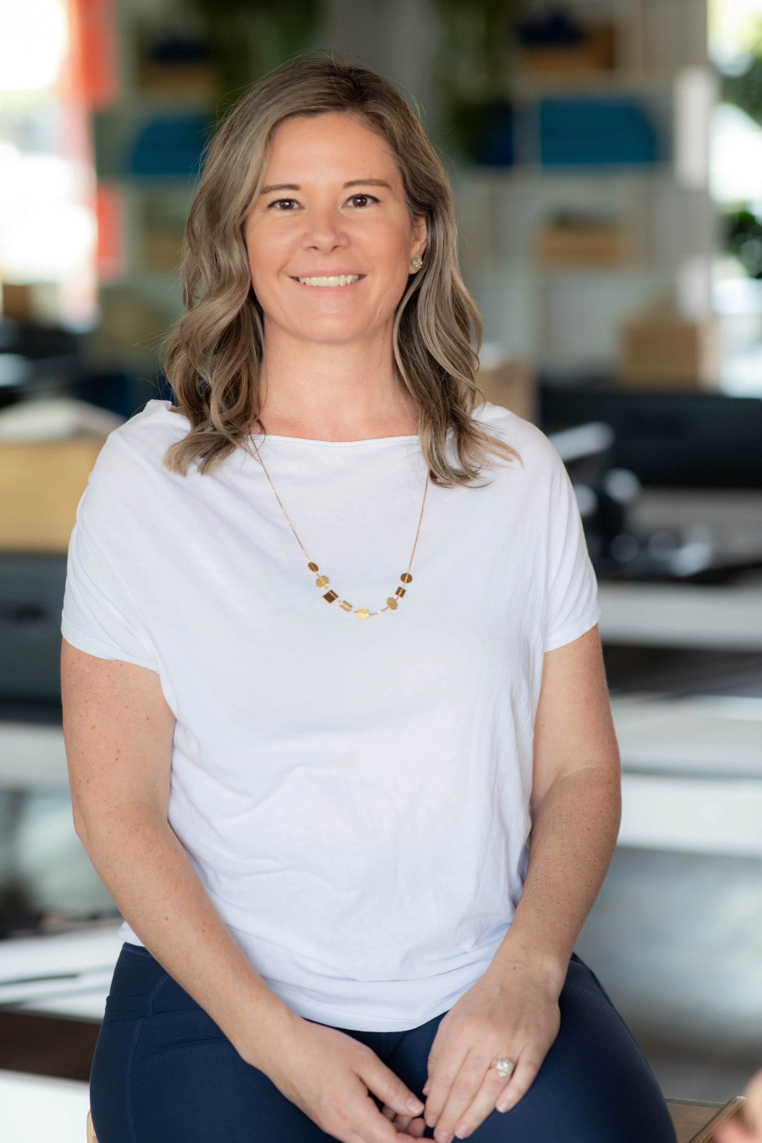 A woman with shoulder-length wavy hair, wearing a white t-shirt and a necklace with geometric shapes, sitting in an office or workspace environment.