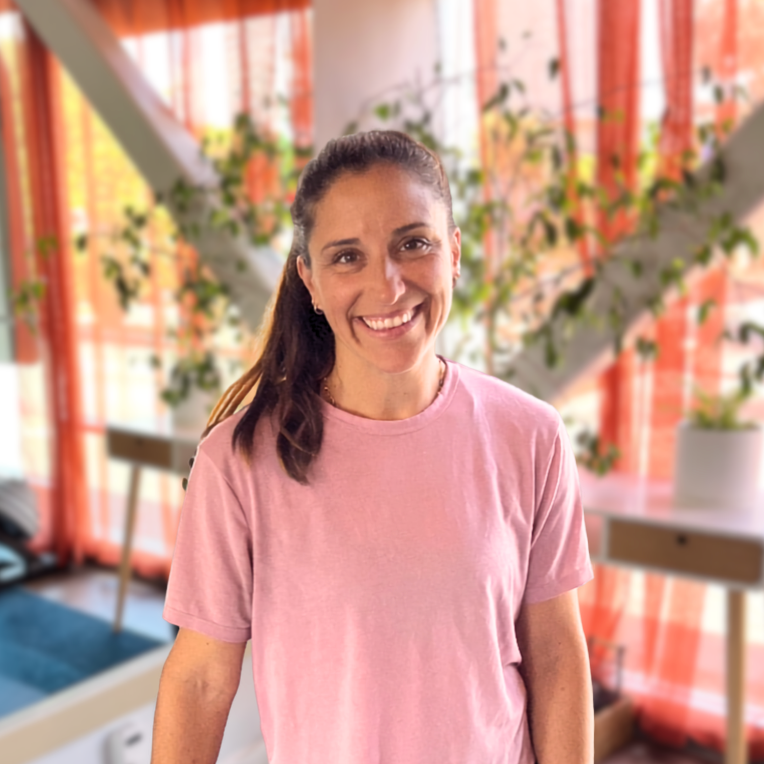 A woman with dark hair in a ponytail, smiling, wearing a pink T-shirt in a brightly lit room with orange curtains, potted plants, a wooden table with a white top, and a chair.