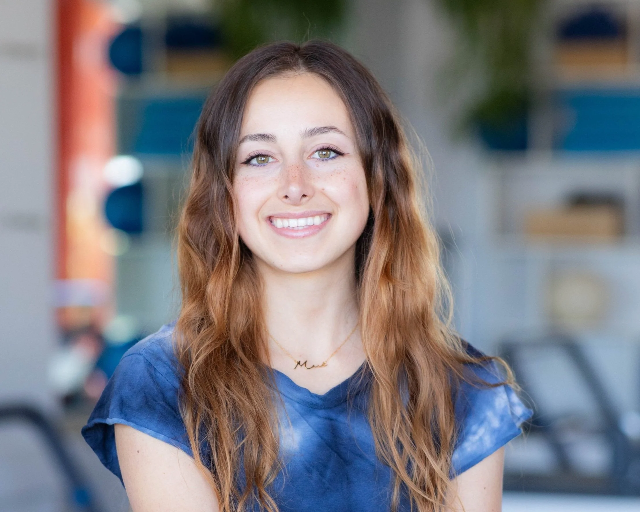 A young woman with long, wavy auburn hair, smiling, wearing a blue tie-dye shirt and a delicate gold necklace with a small name pendant, standing outdoors with blurred background.