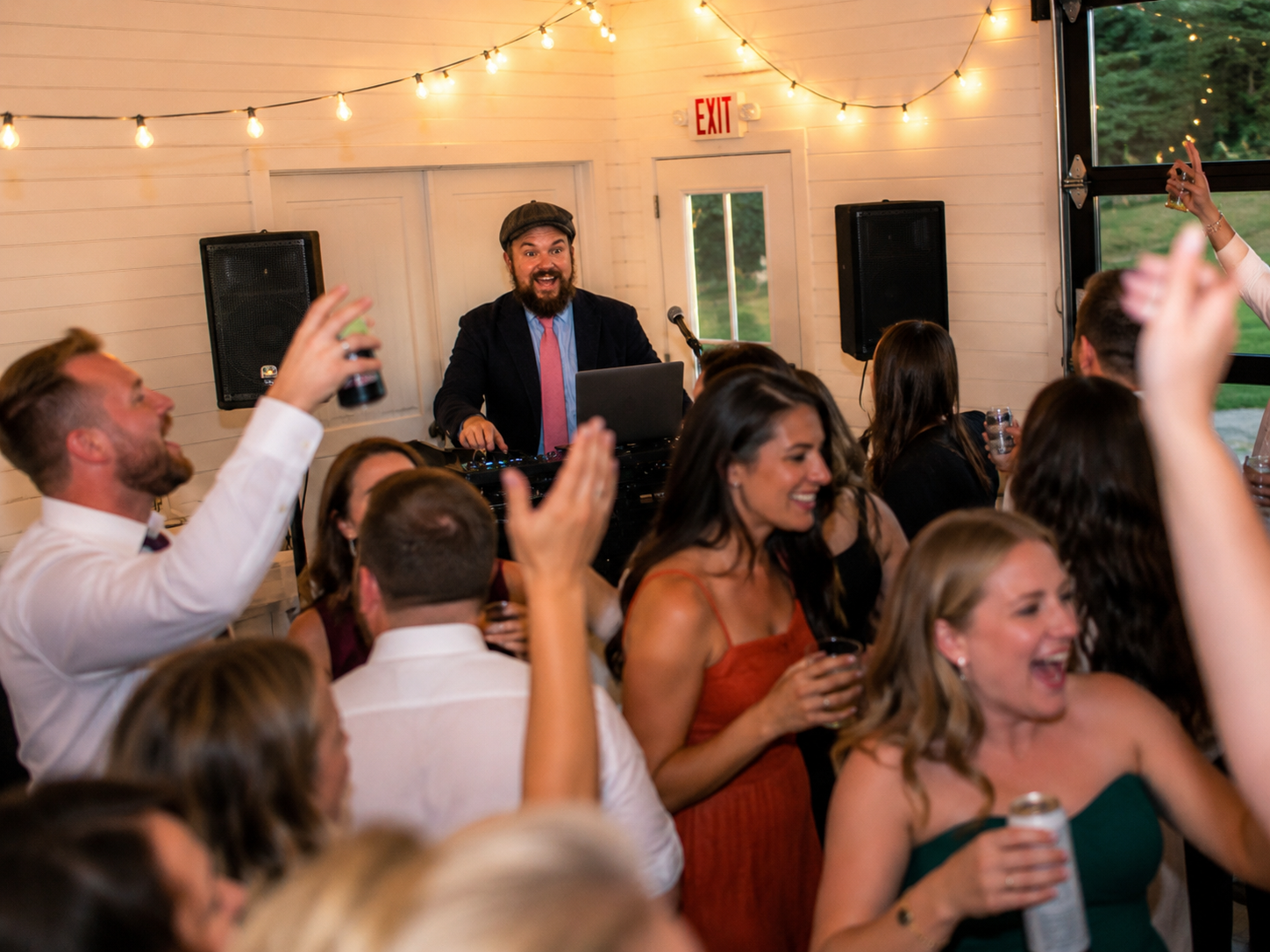 People at a party dancing and enjoying music with a DJ playing in the background under string lights.