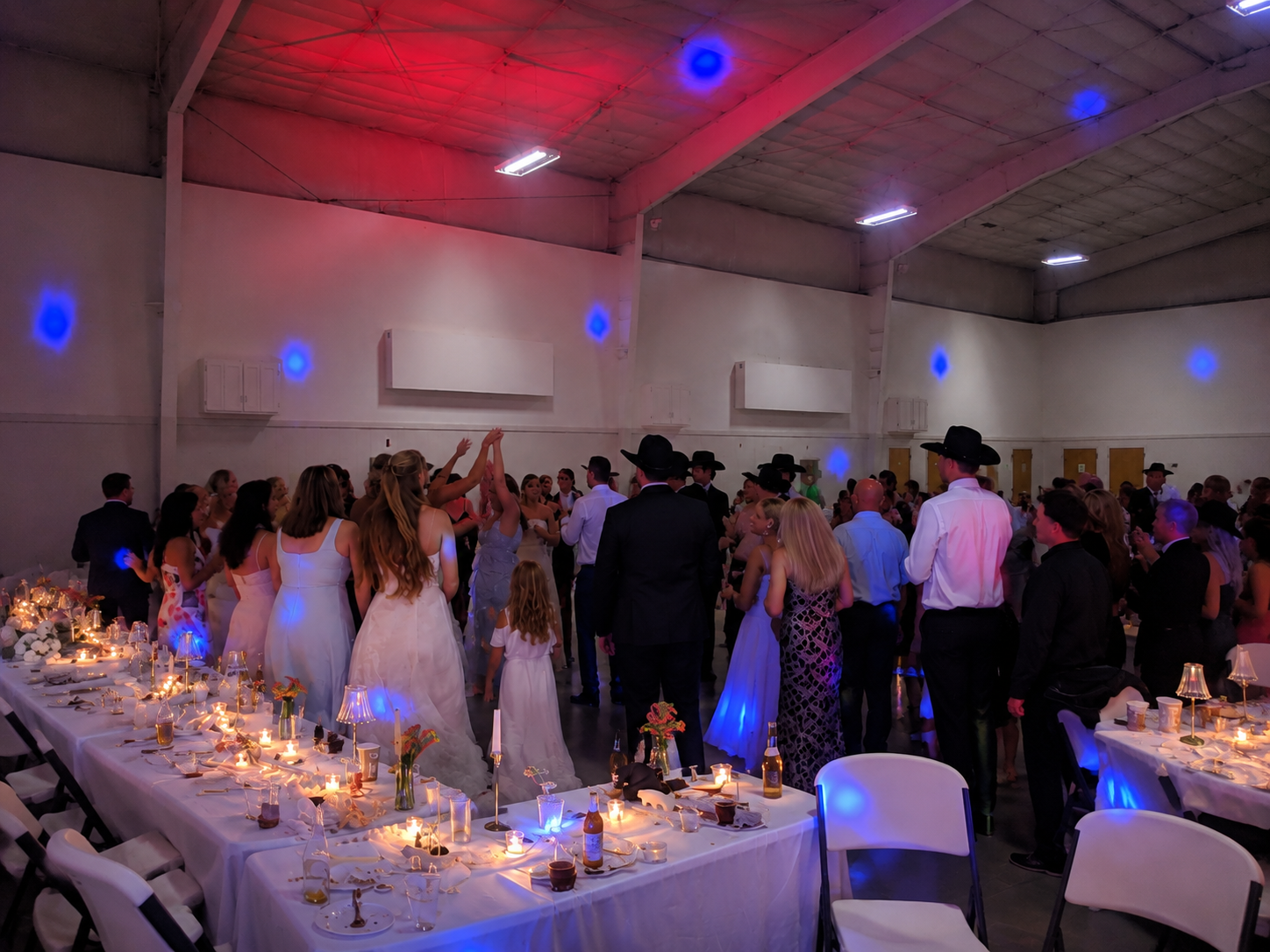 A lively party or celebration in a large indoor venue with people dancing, some wearing formal attire and cowboy hats. The foreground features a long table decorated with candles, small vases, and drinks. The room has high ceilings with colorful ligh