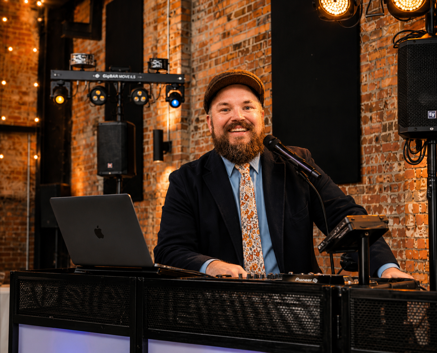 A man with a beard, wearing a brown hat, dark blazer, light blue shirt, and floral tie, smiling at a microphone in a brick-walled venue with stage lights and audio equipment. He is seated at a DJ setup with a laptop and mixer.