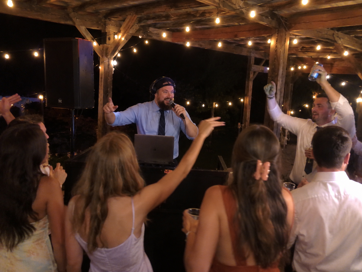 People dancing and celebrating at a party under string lights, with a DJ wearing headphones and holding a microphone.