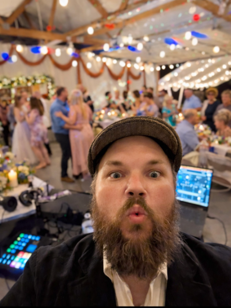 DJ Jazzy Josh reacting during a lively wedding reception with guests dancing, warm string lighting, and an active dance floor in the background.