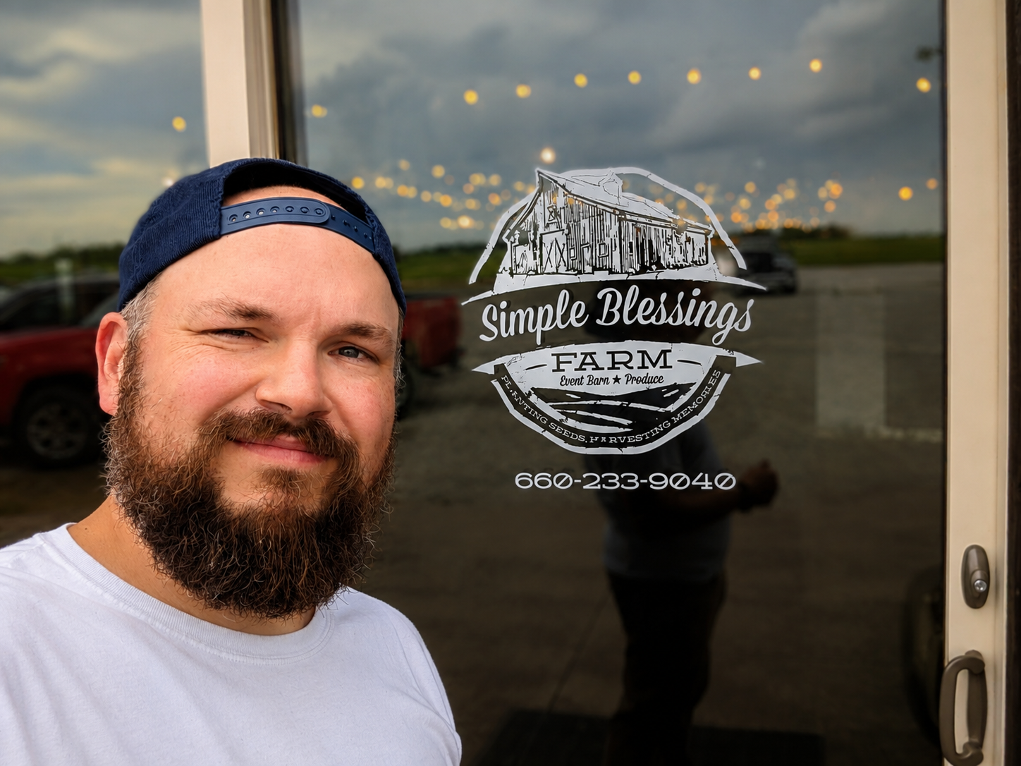 Man with a beard and blue cap standing in front of a glass door with Simple Blessings FARM logo and phone number, outdoors during evening with cloudy sky and parked vehicles reflected on the glass.