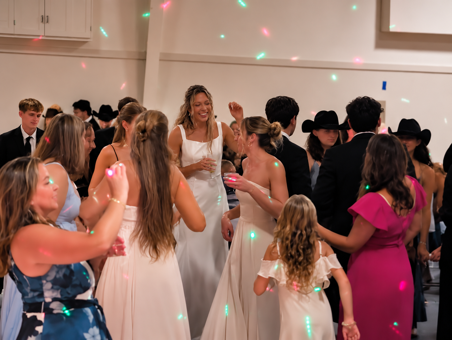 Group of people dancing and socializing at a party, dressed in formal and semi-formal attire, with colorful lights.