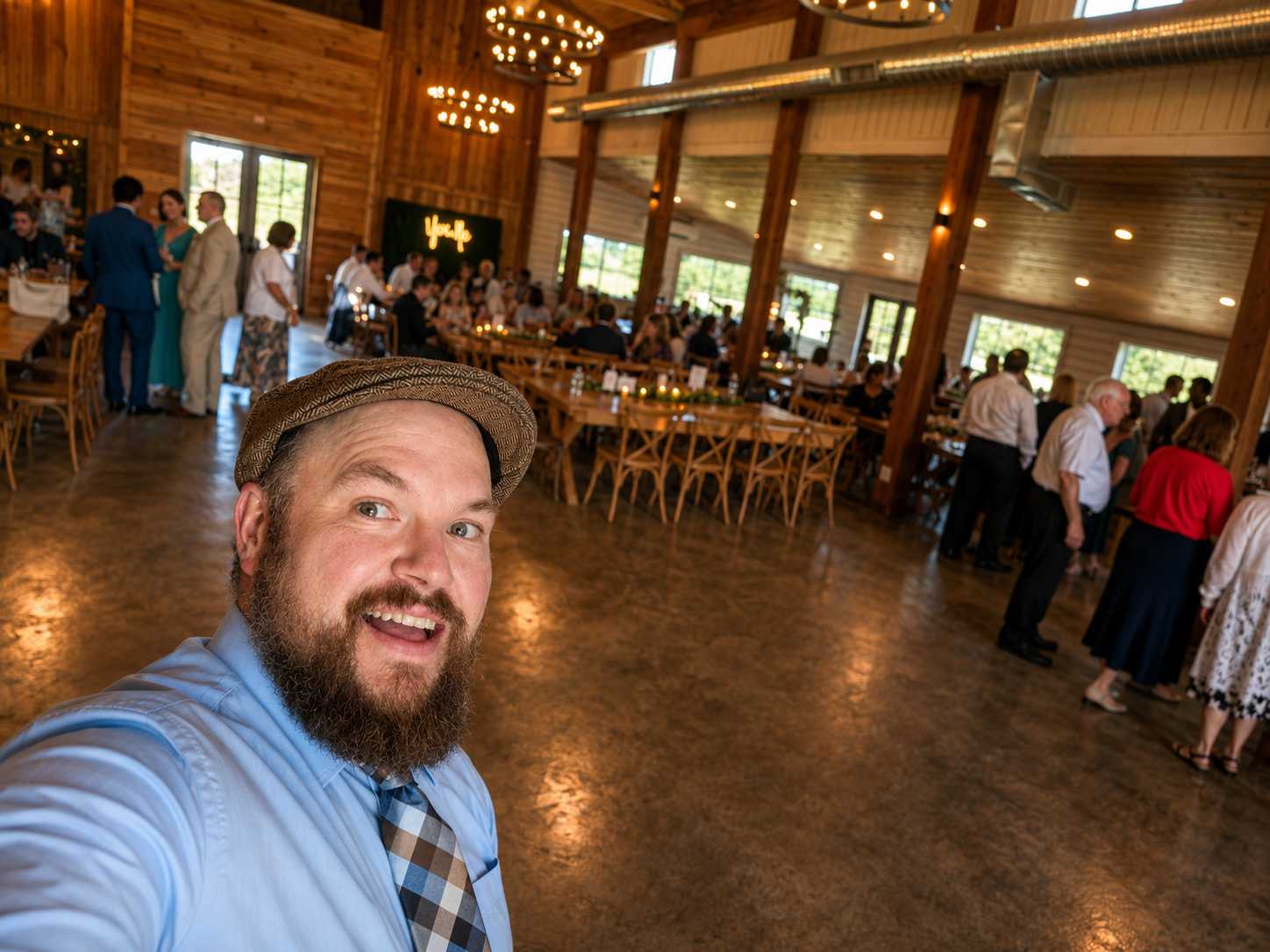 A bearded man in a blue shirt and patterned tie taking a selfie at a large wooden event hall filled with people, some of whom are standing in line, and others are sitting at long tables decorated for a celebration.