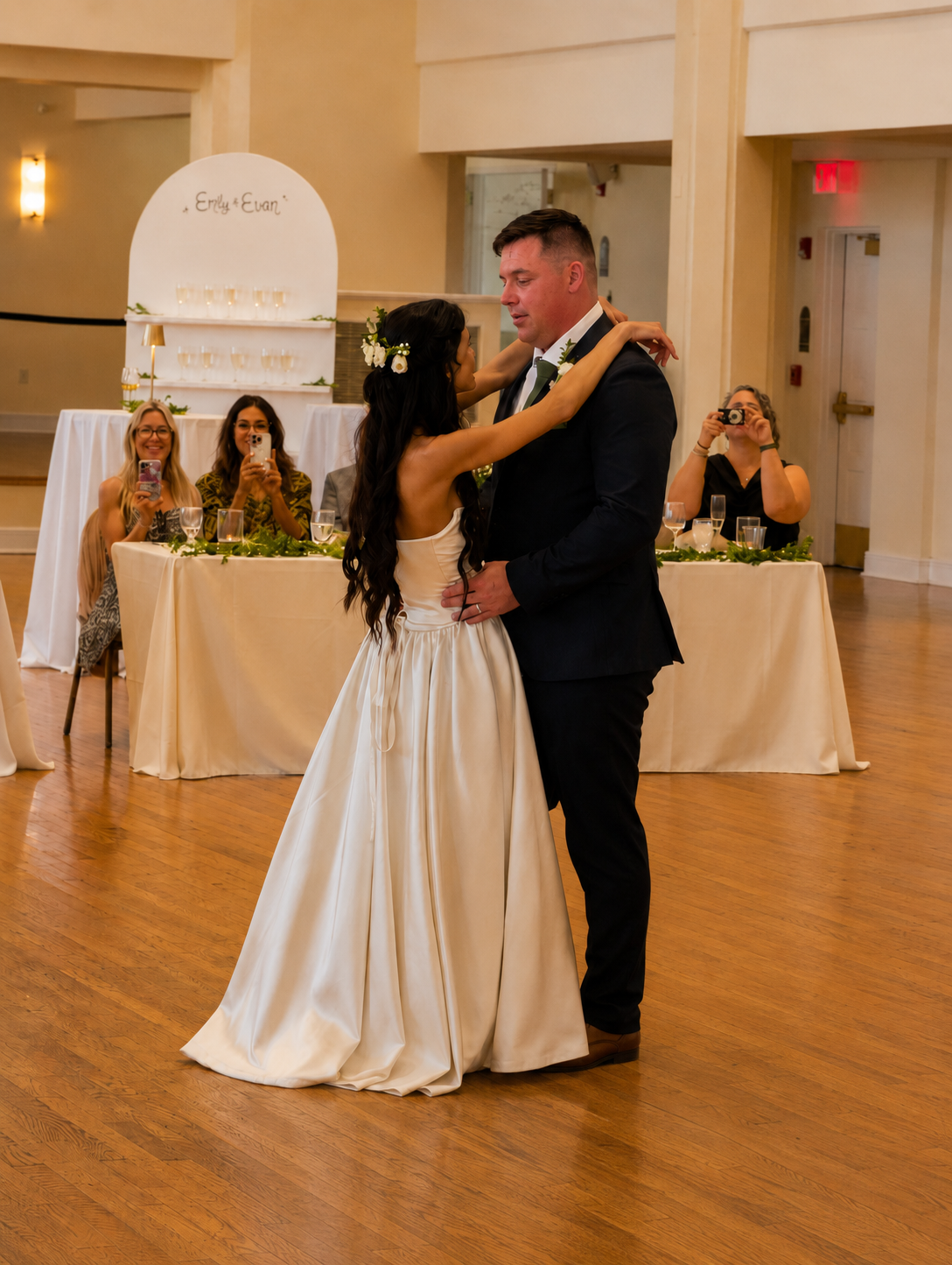 A bride and groom share a dance at their wedding reception, with three women at a table behind them taking pictures and smiling.