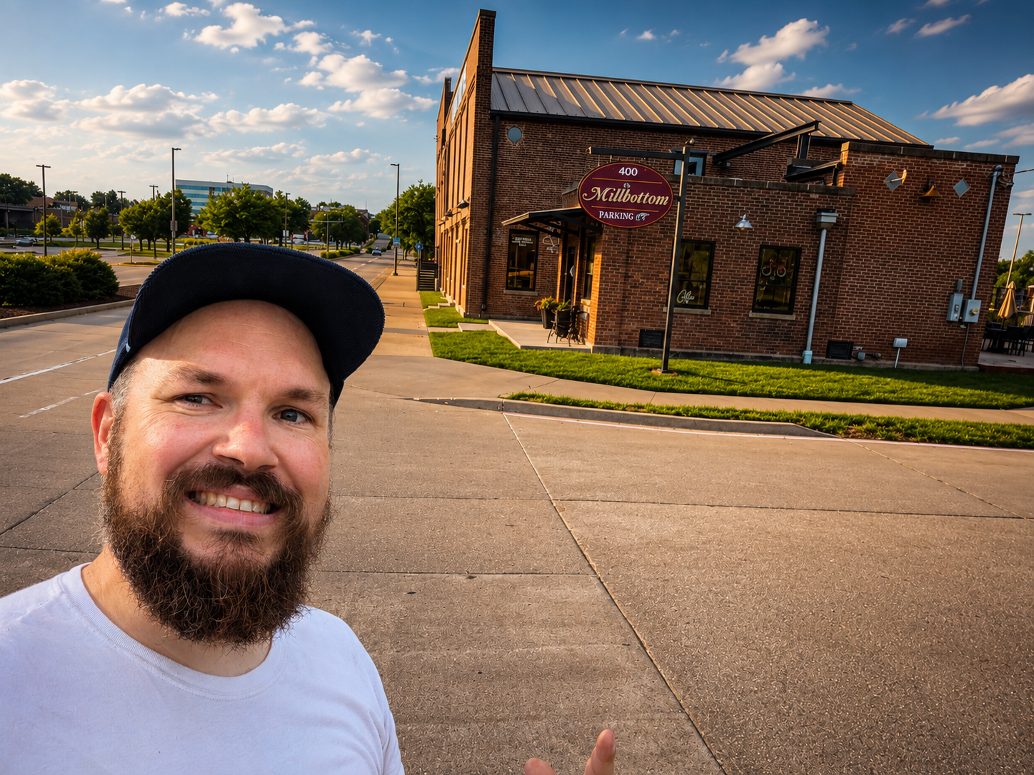 A bearded man smiling and pointing at a brick building with a sign that says 'Mulbottom Parking' under a blue sky with scattered clouds