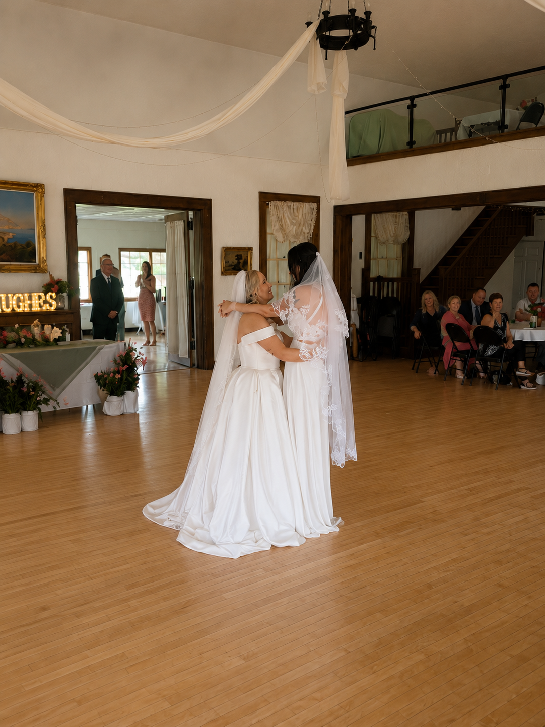 Two brides in wedding dresses sharing a dance at a wedding reception, with guests watching and standing in the background.