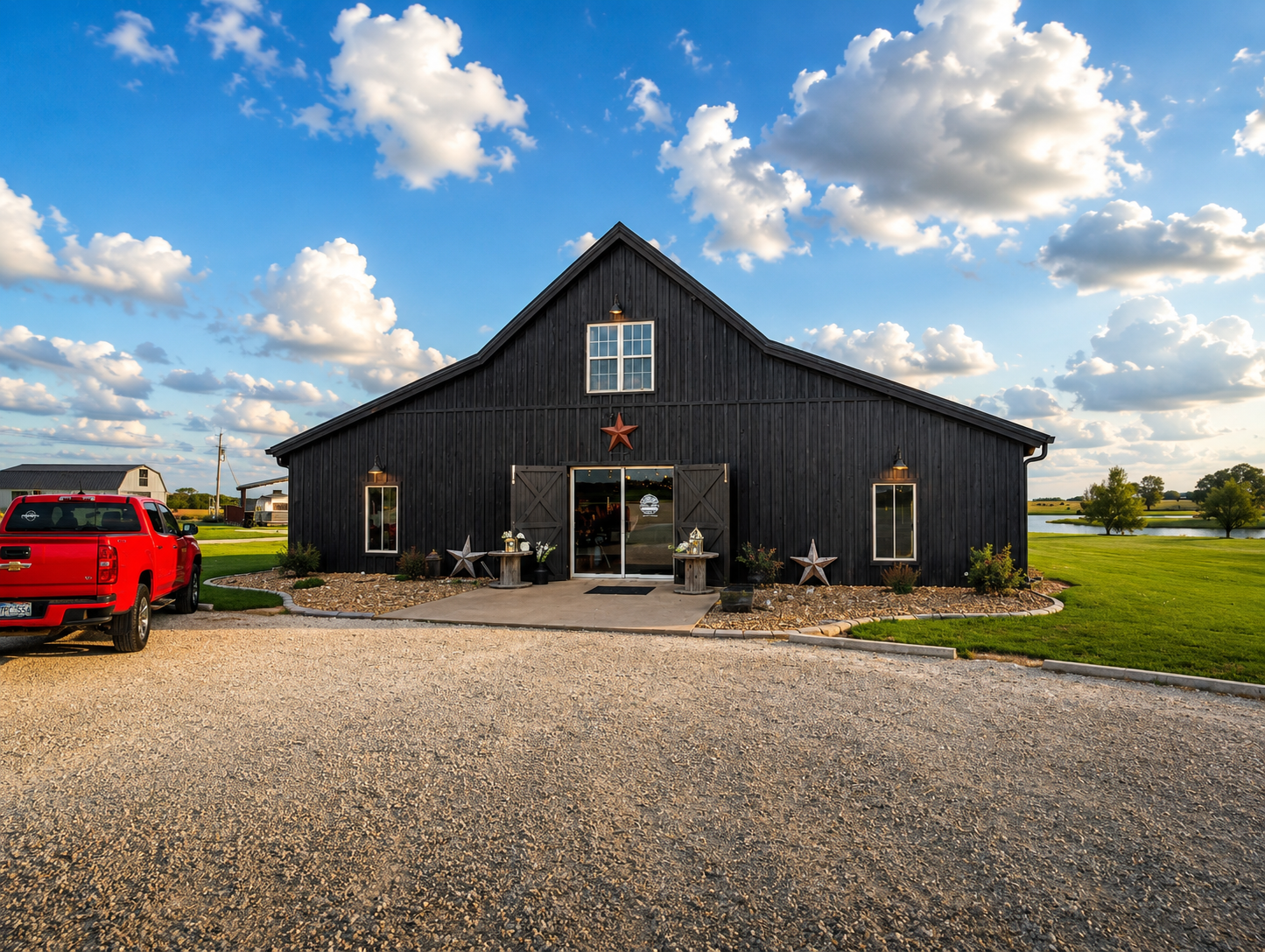 A large black barn-style building with open wooden doors decorated with star-shaped ornaments, two pairs of windows, outdoor lighting, and a porch. A red pickup truck is parked on a gravel driveway. The sky is blue with fluffy clouds, and there are g
