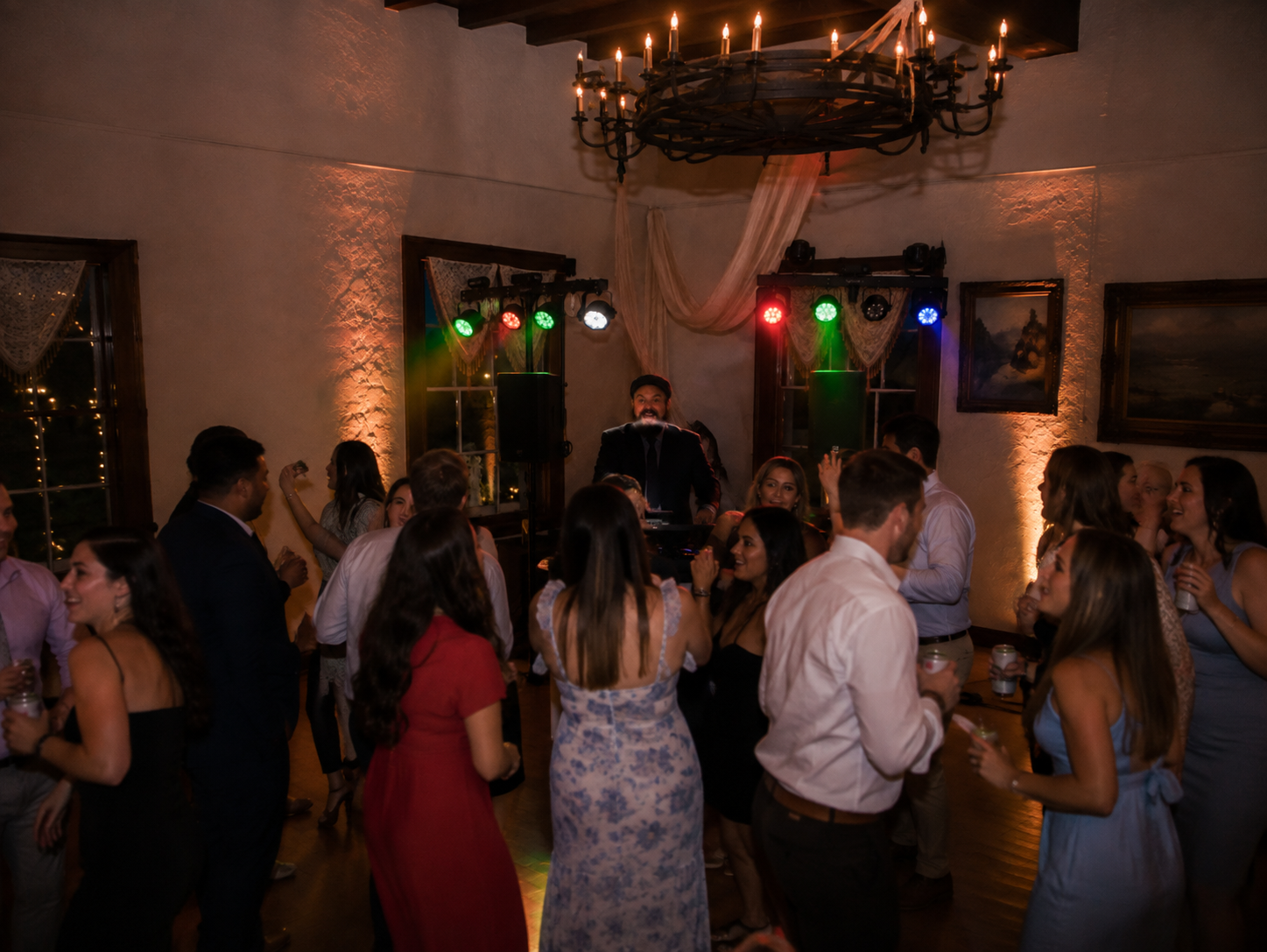 People dancing and socializing at a party with a DJ or band playing music, colorful stage lights, and warm ambient lighting in a decorated indoor venue.