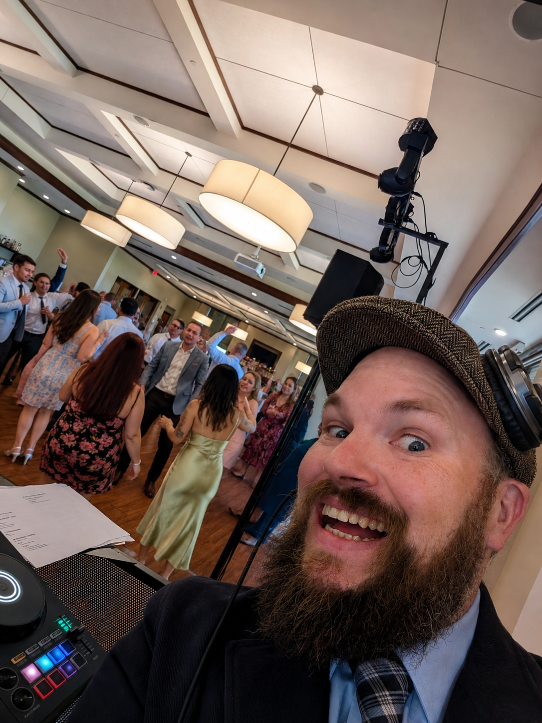 DJ Jazzy Josh smiling behind the DJ booth during a packed wedding reception with guests dancing under elegant ballroom lighting.