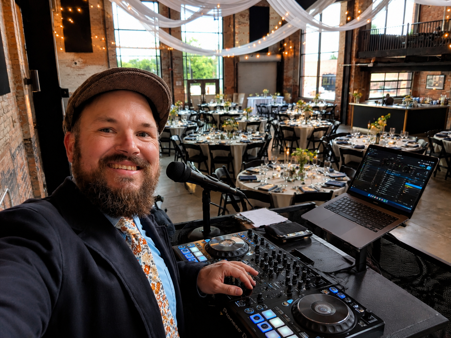 A bearded man in a hat smiling at a camera in a decorated event space with round tables, floral centerpieces, and string lights, standing behind DJ equipment, a laptop, and a microphone.