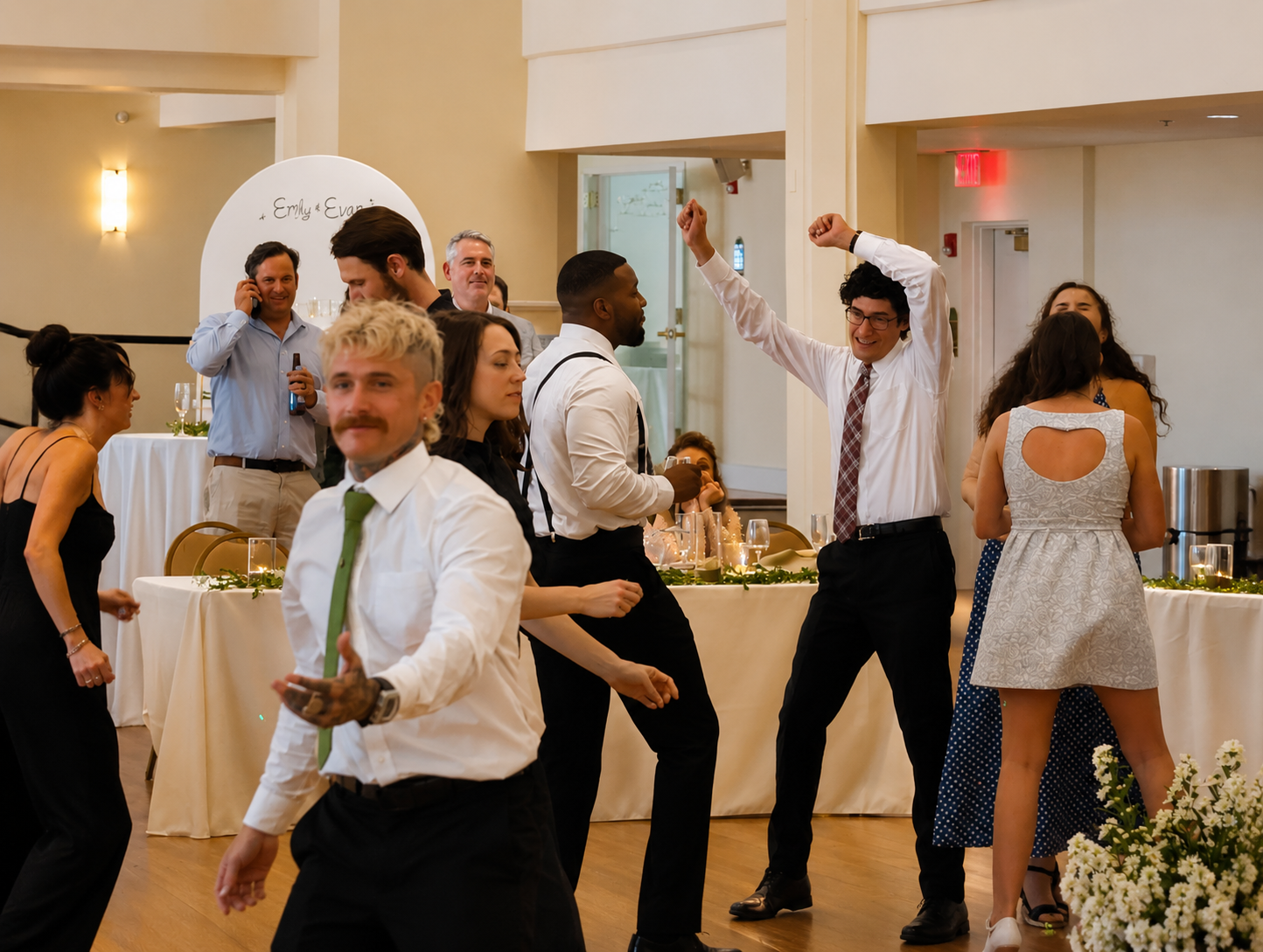 People dancing and socializing at an indoor celebration or wedding reception, with a decorated table, floral arrangements, and a guest looking at the camera.