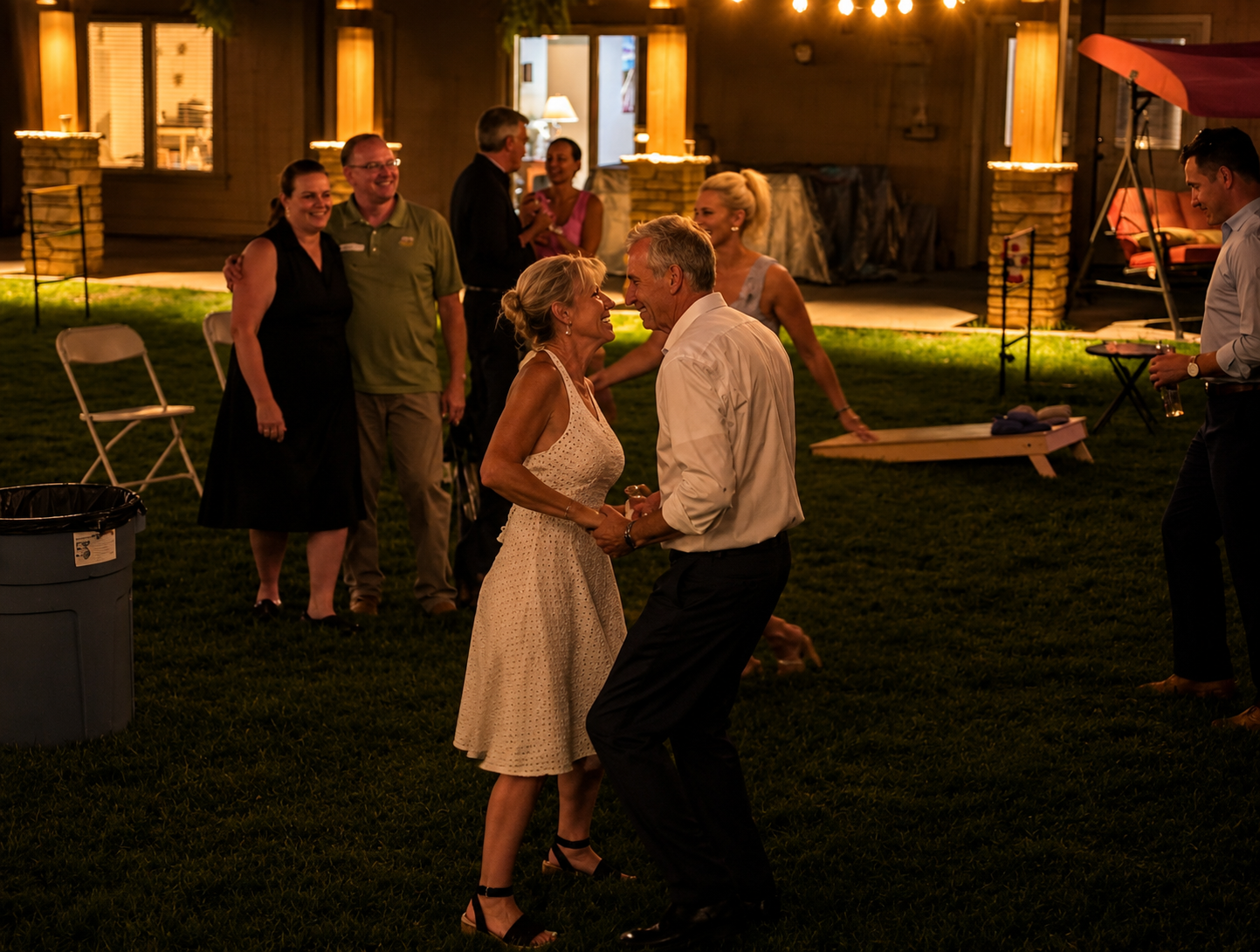 A couple dancing and smiling at a nighttime outdoor event with other people socializing in the background, illuminated by warm lighting.