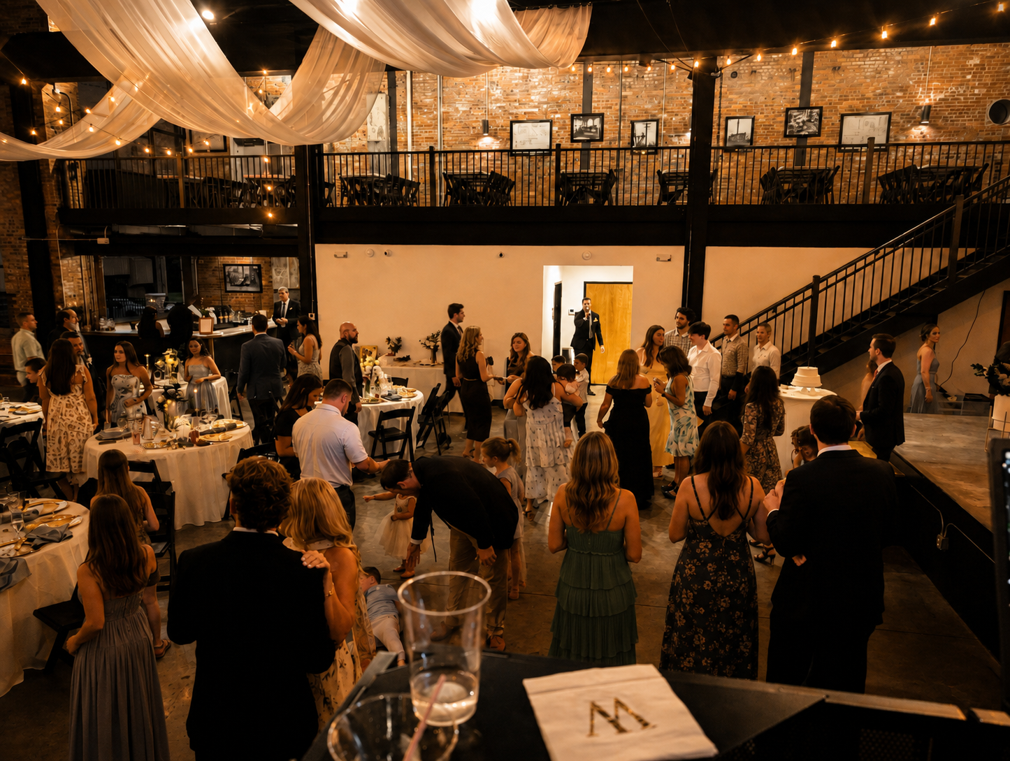 Indoor event at a rustic-style venue with exposed brick walls, string lights, and draped fabric ceiling, featuring guests gathering around tables and a dance floor, celebrating a wedding or similar occasion.