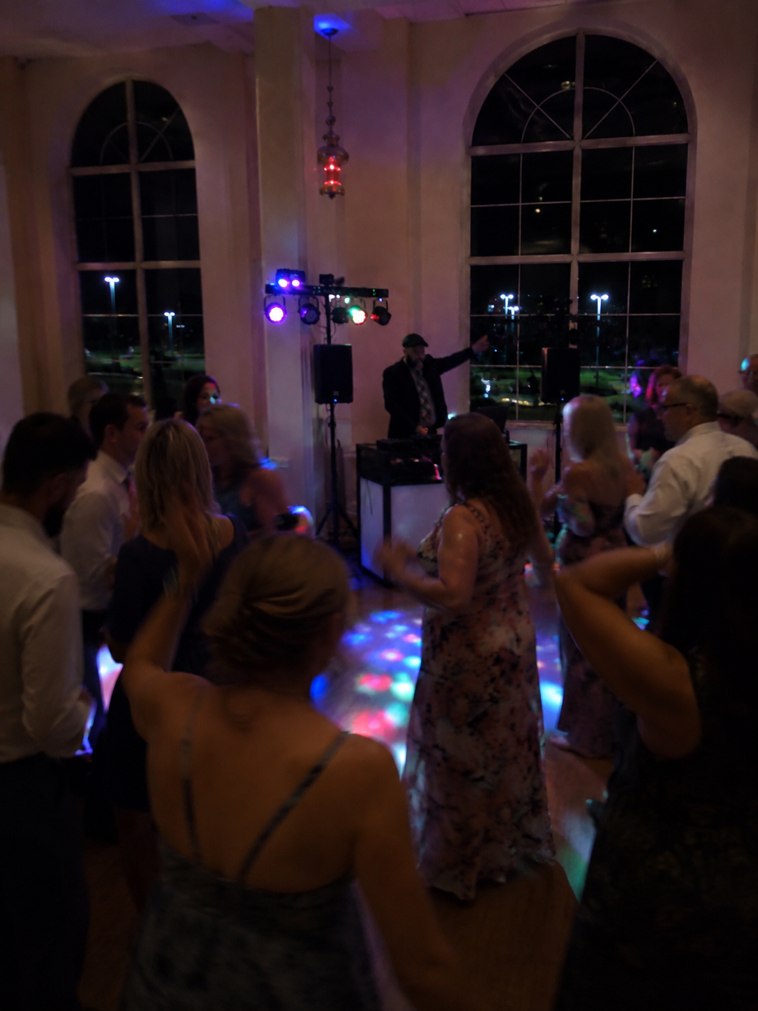 People dancing and socializing at a party with a DJ playing music, colorful lights, and large windows showing a dark outdoor scene.
