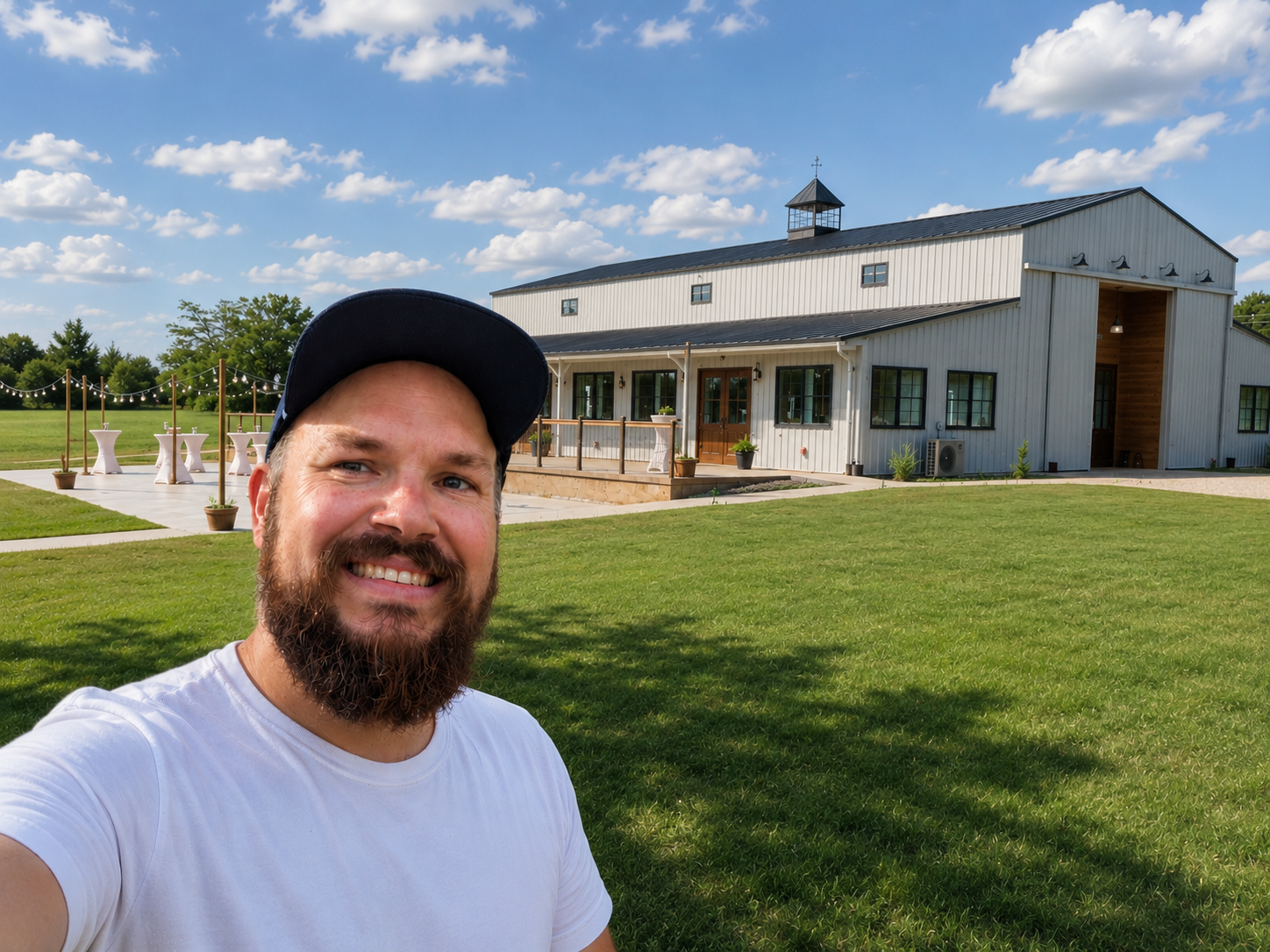 A man with a beard smiling in the foreground, taking a selfie in front of a modern barn-style building on a sunny day with a blue sky and clouds, surrounded by green grass and trees.