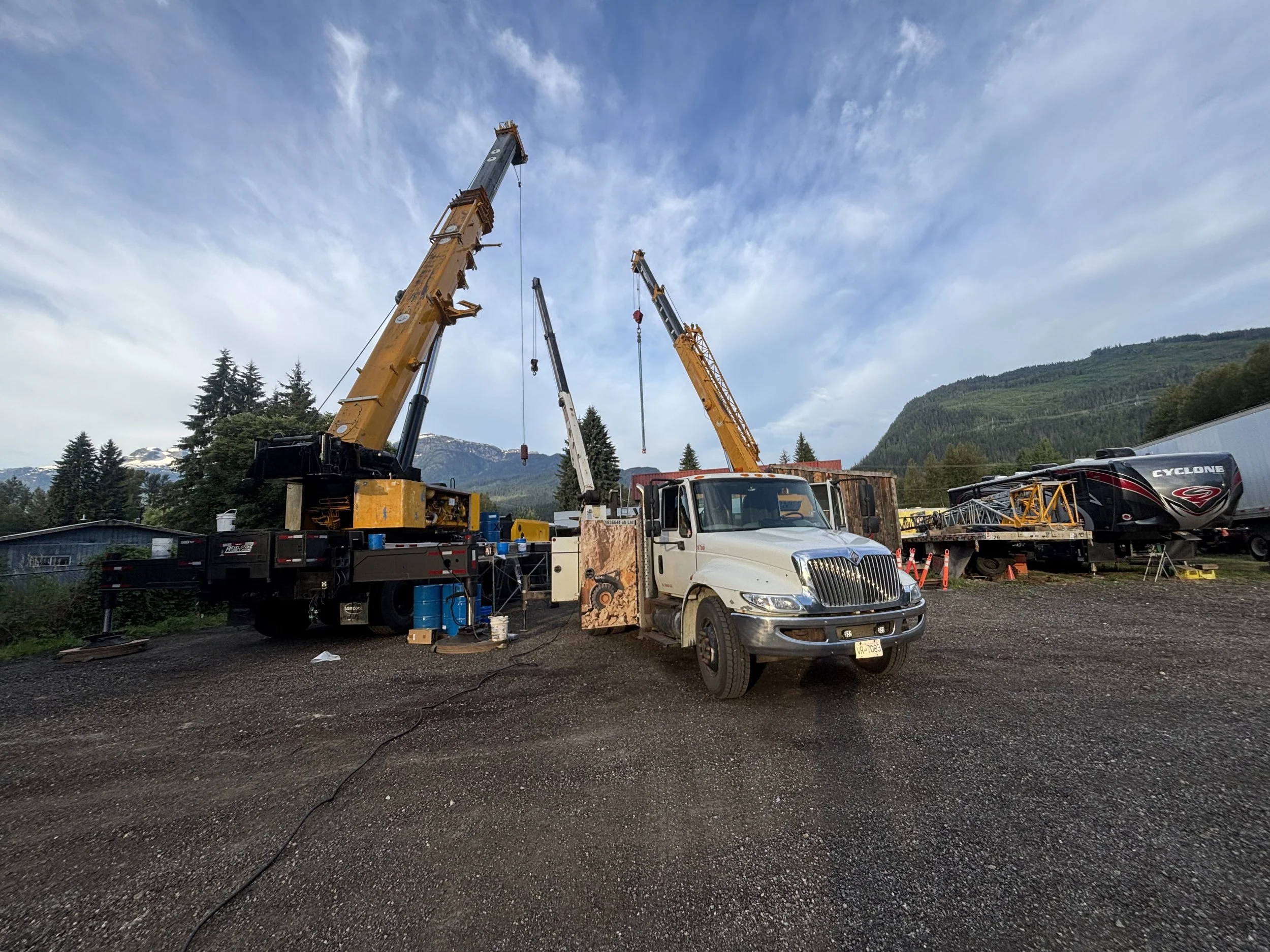 Construction site with multiple cranes lifting objects, a truck, and a recreational vehicle against mountainous scenery with trees and a partly cloudy sky.
