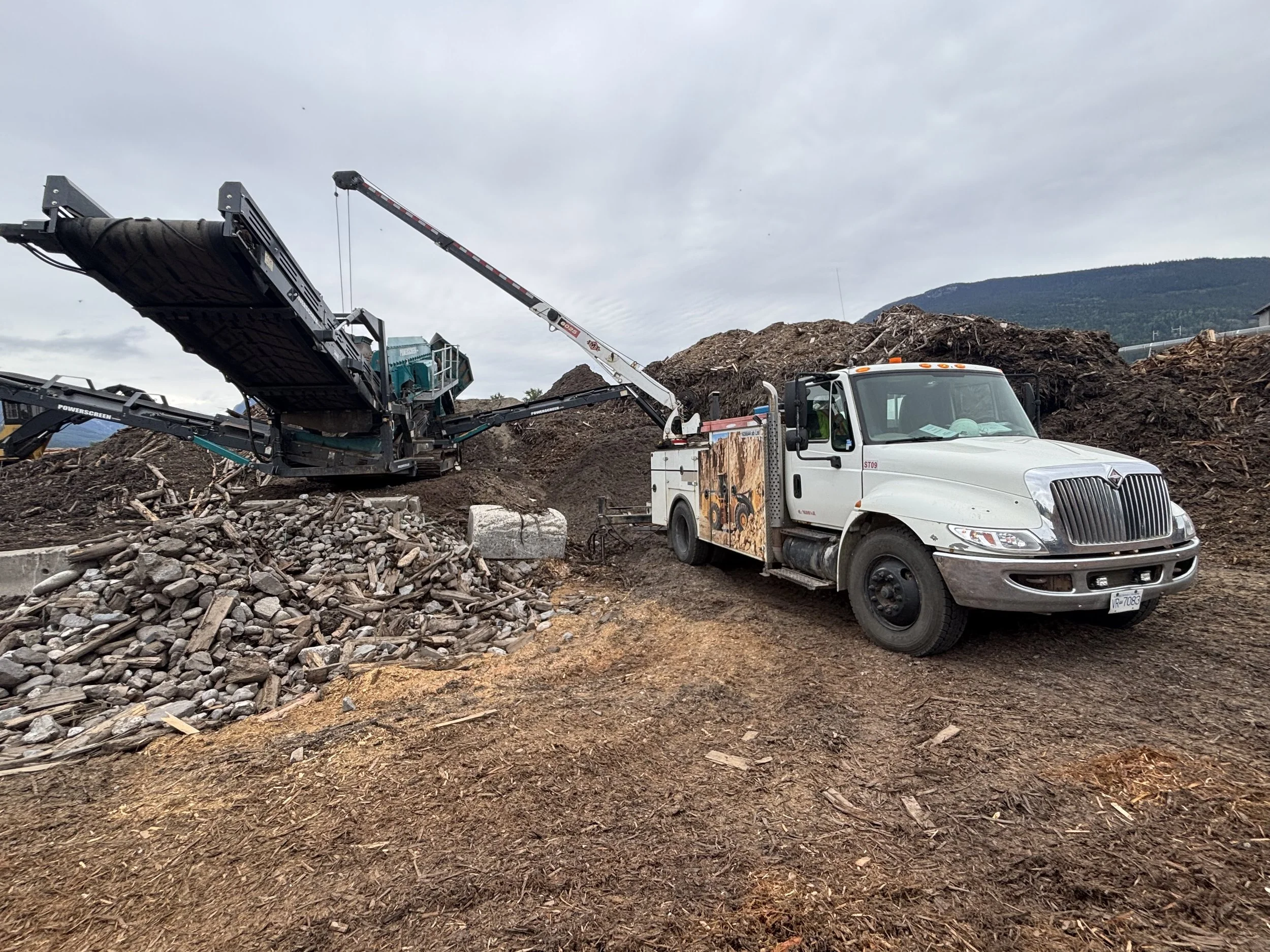 Construction site with a large machine and a truck on dirt ground, mountains in the background, overcast sky.