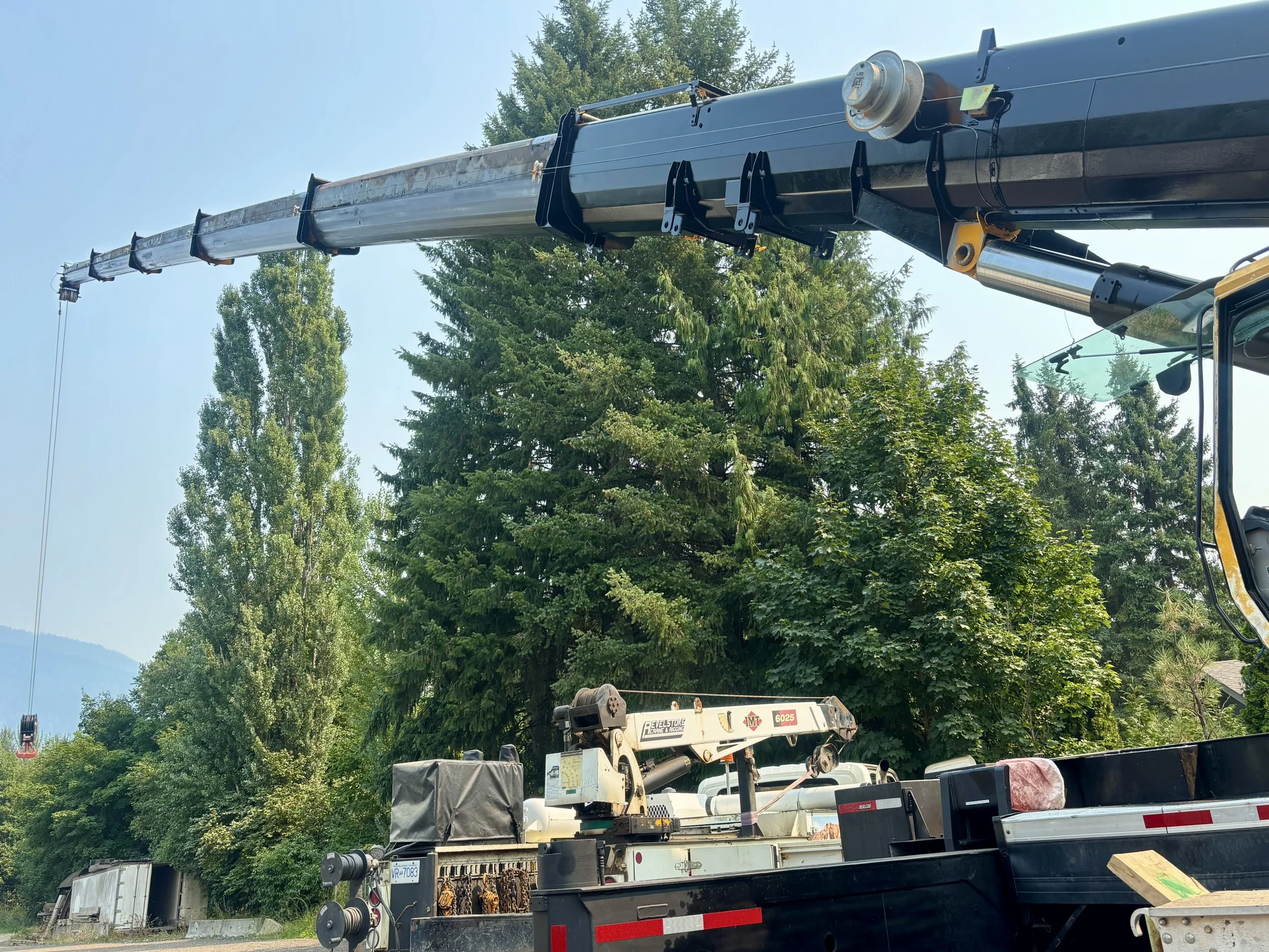 A construction crane lifting a long metallic beam with trees and a clear blue sky in the background.