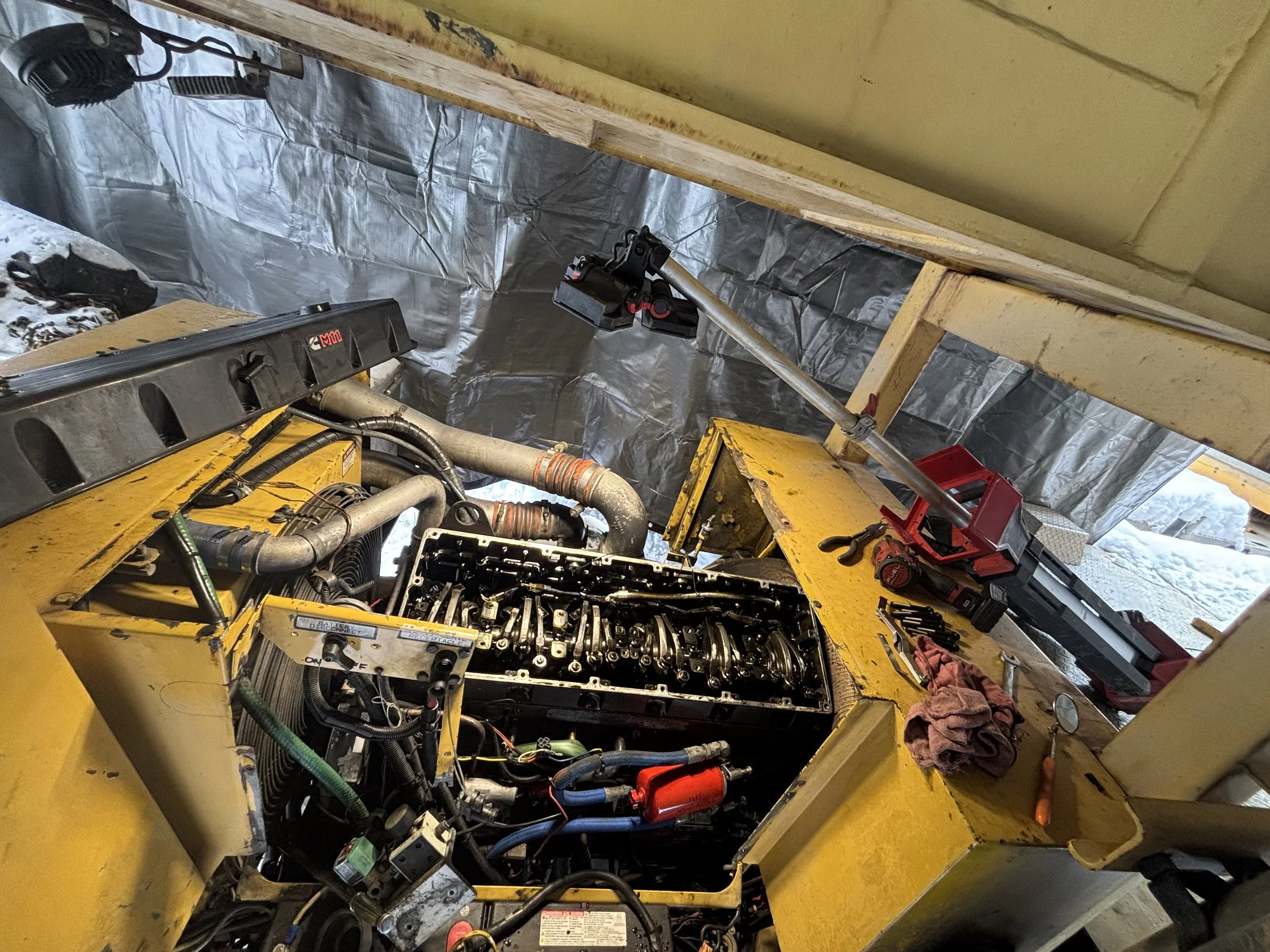 The image shows the inside of a construction or repair site with yellow metal equipment, tools, and wires. Part of an engine or machinery is visible, and there are metallic insulation panels on the walls. The ceiling is made of brick, and there are some red and black tools placed on the yellow machinery.