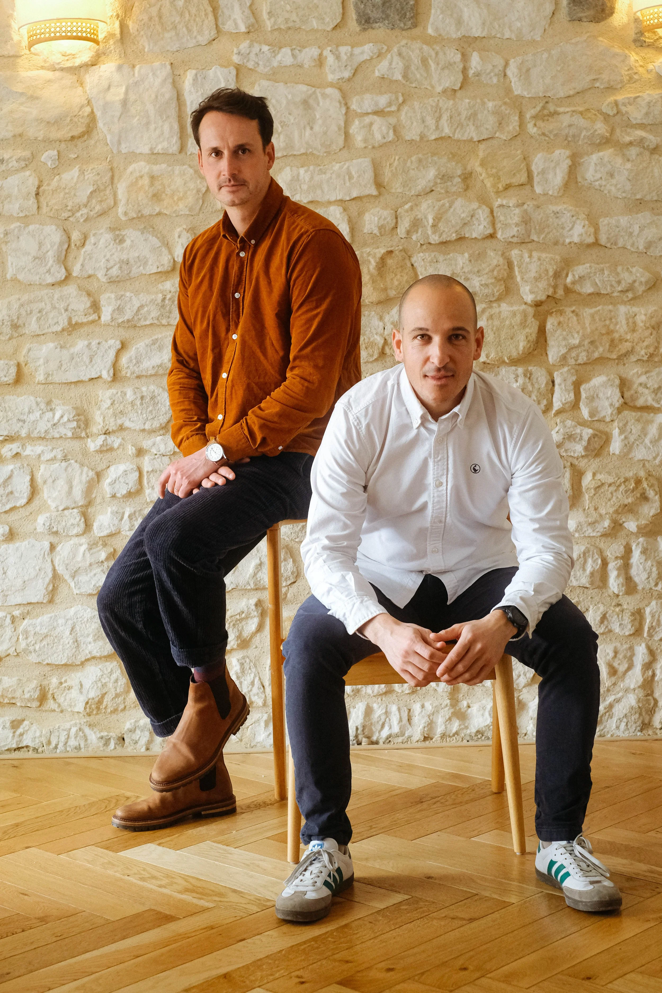 Deux hommes posent dans une pièce avec un mur en pierre blanche en arrière-plan. L'homme à gauche est vêtu d'une chemise marron et de bottes en cuir, assis sur un tabouret, tandis que l'homme à droite porte une chemise blanche et des sneakers, assis sur une chaise en bois. Les deux ont une expression sérieuse.